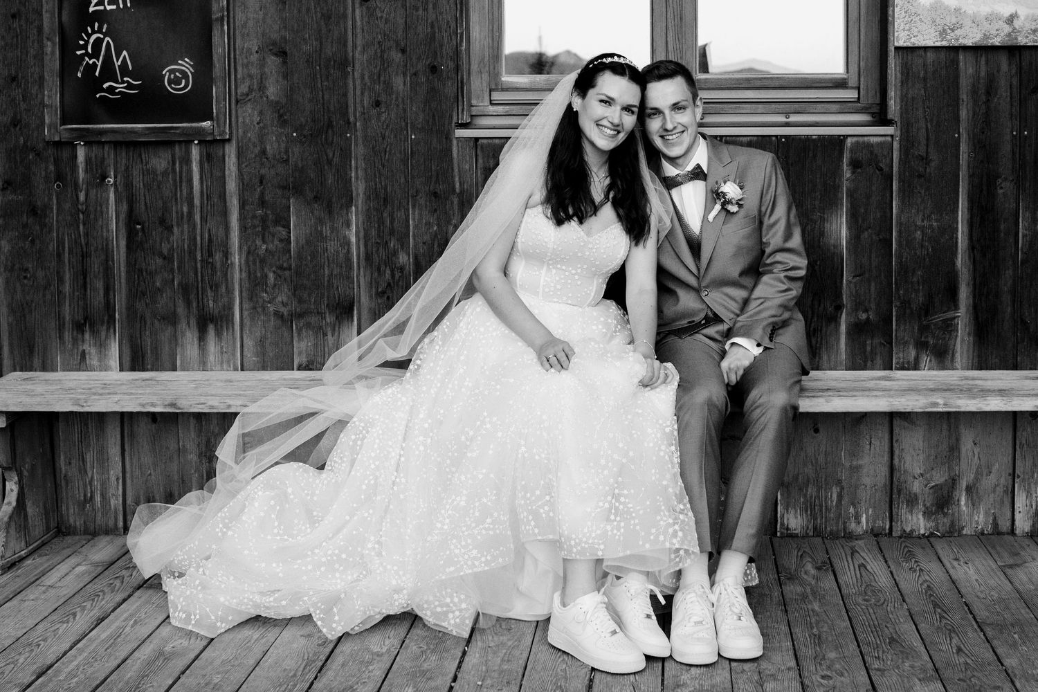 Black and white photo of a bride in a white gown and groom in a suit sitting together on a wooden porch.