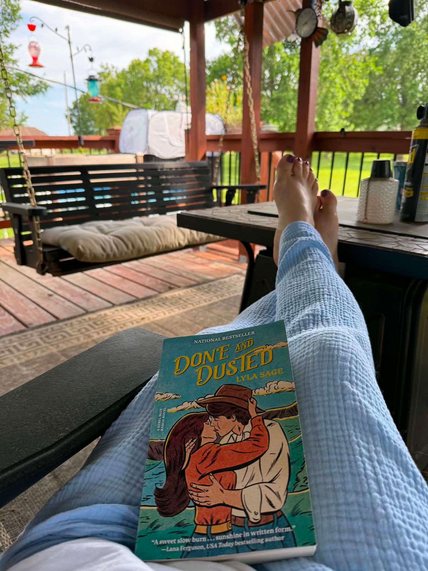 Person relaxing on a deck reading 'Done and Dusted' novel, with outdoor patio furniture and greenery in the background.