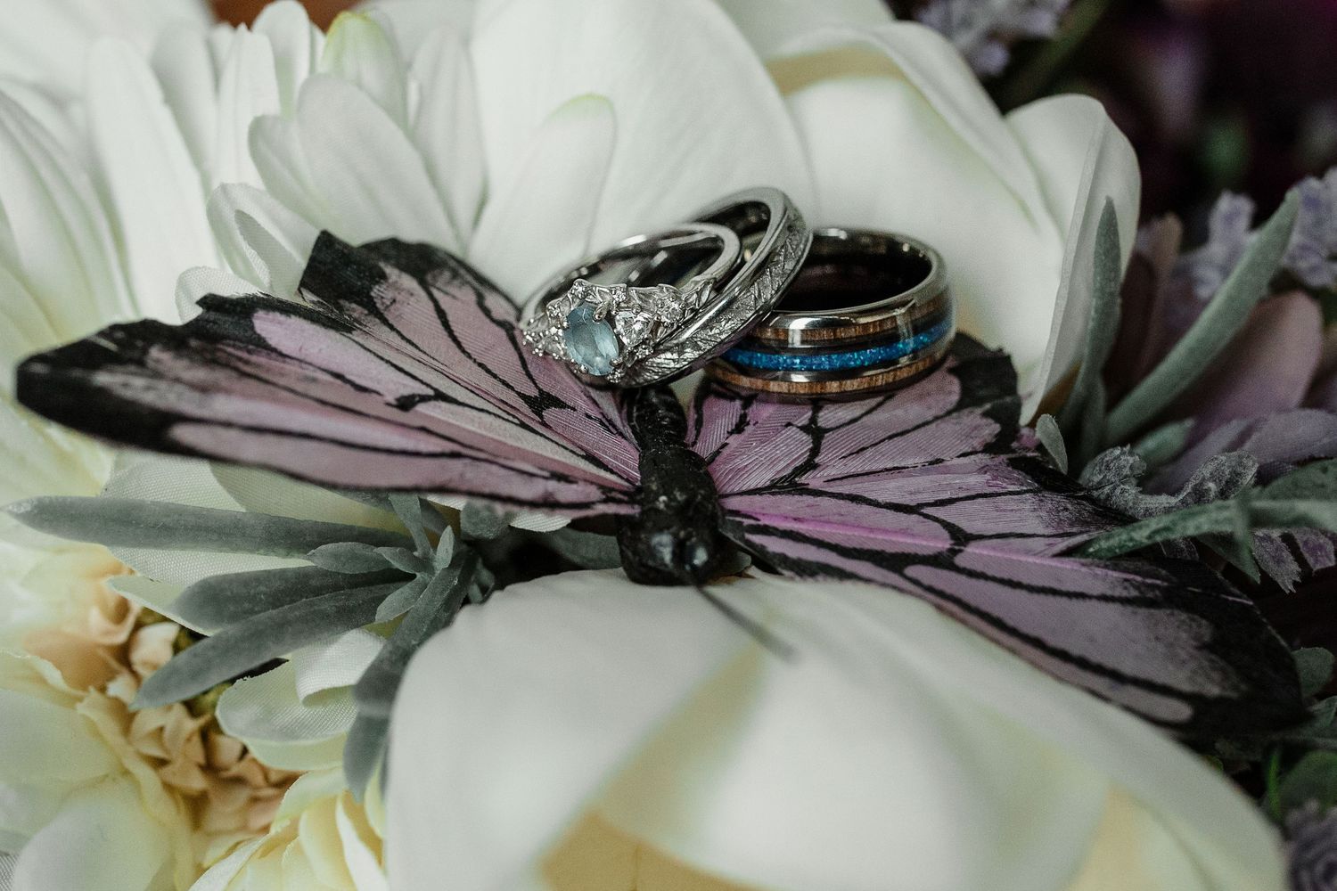 Wedding rings resting on a decorative butterfly among white flowers, featuring diamond, blue opal, and black bands.