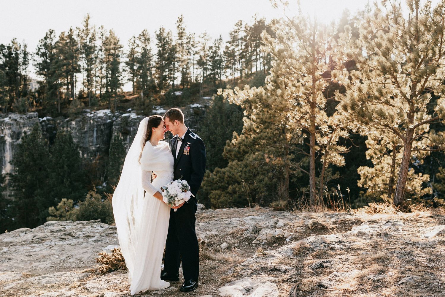 Bride and groom share a romantic kiss on rocky cliffside surrounded by pine trees in stunning outdoor wedding photo.