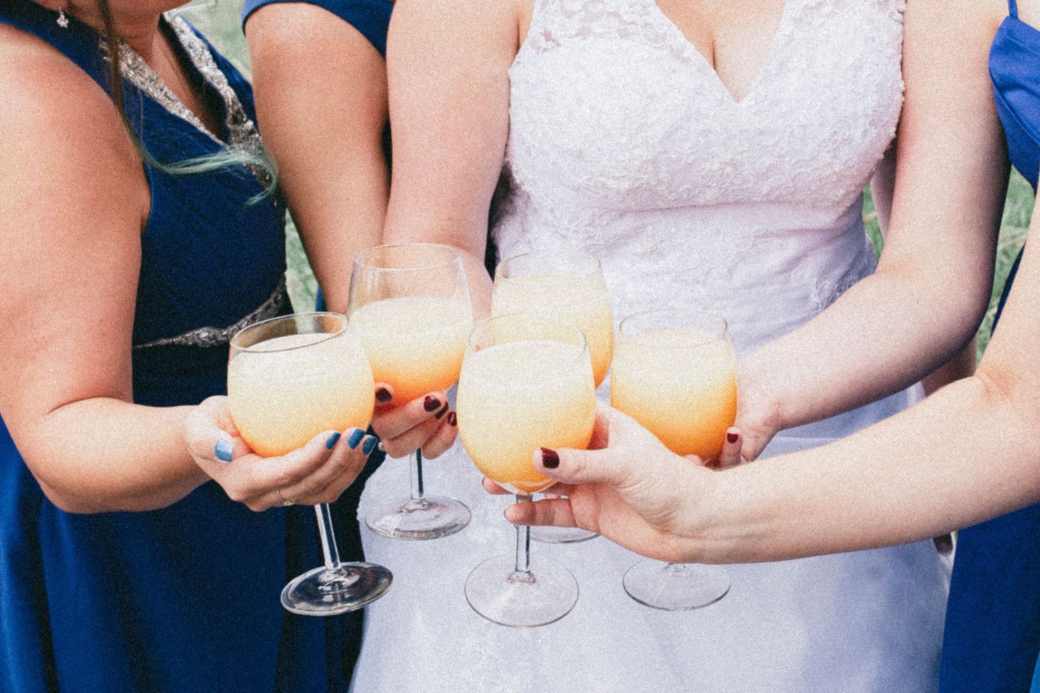 Bride and bridesmaids in blue dresses toasting with orange cocktails in wine glasses at a wedding celebration.