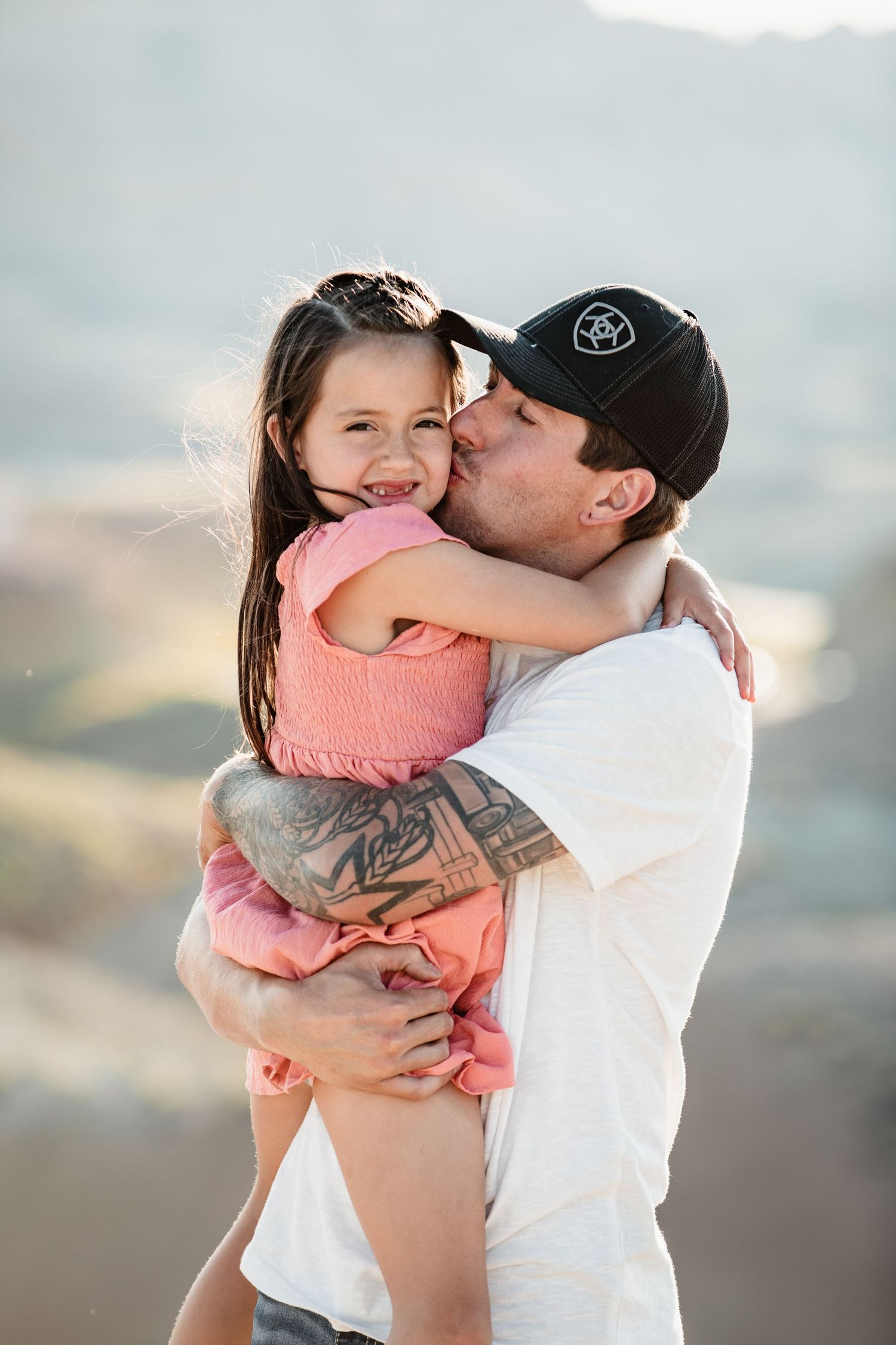 A tattooed man in a black cap kisses a smiling little girl in a pink dress outdoors.