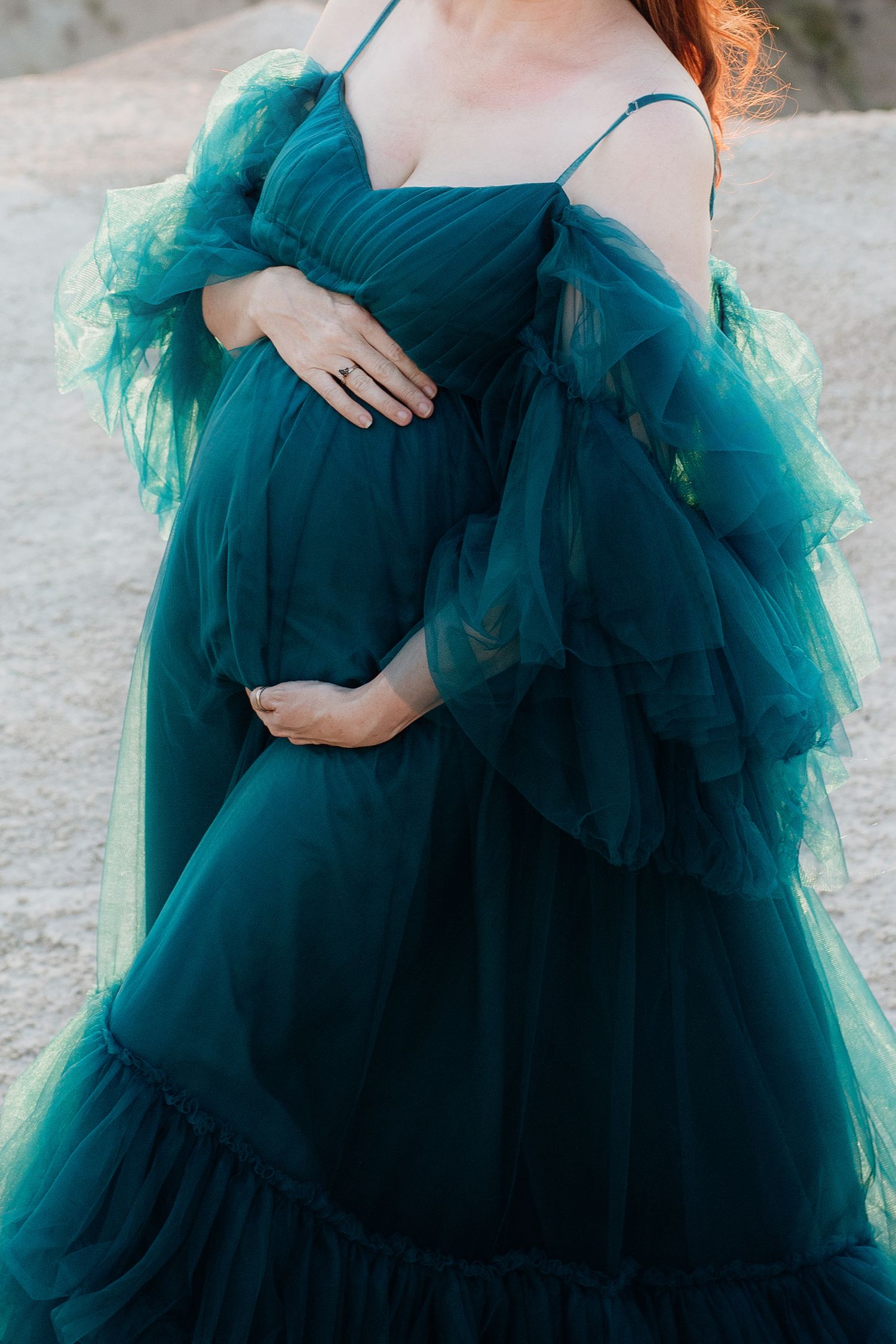 Close-up of pregnant woman in teal tulle maternity gown gently holding her baby bump.
