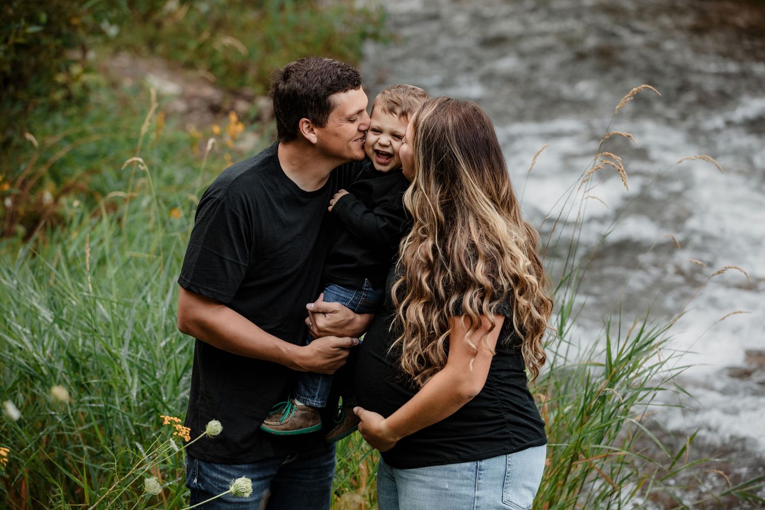 Happy family with toddler laughing as parents kiss child on cheeks near a flowing river outdoors.