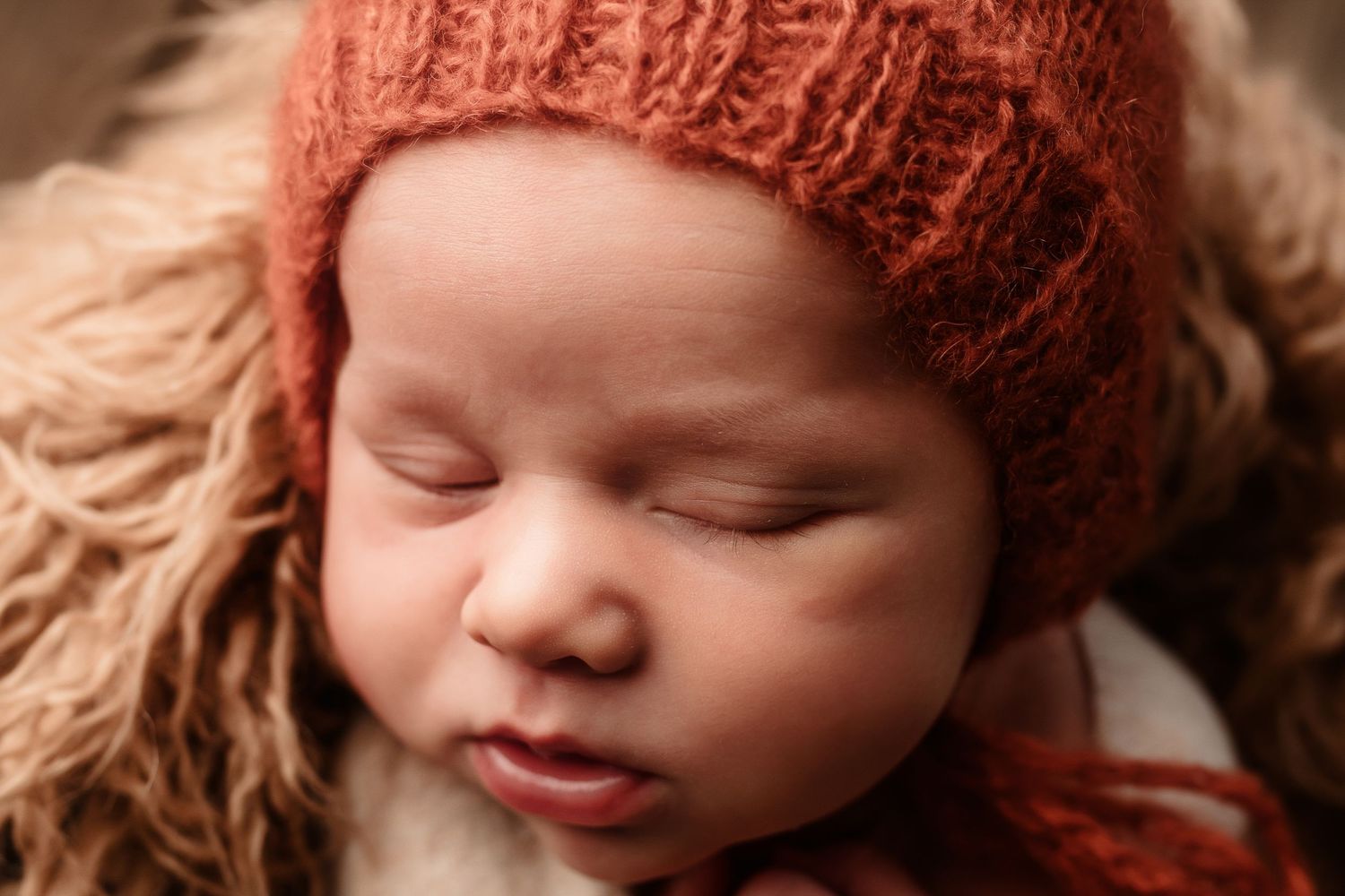 A sleeping baby wearing a knitted orange beanie lies peacefully on soft brown fur.