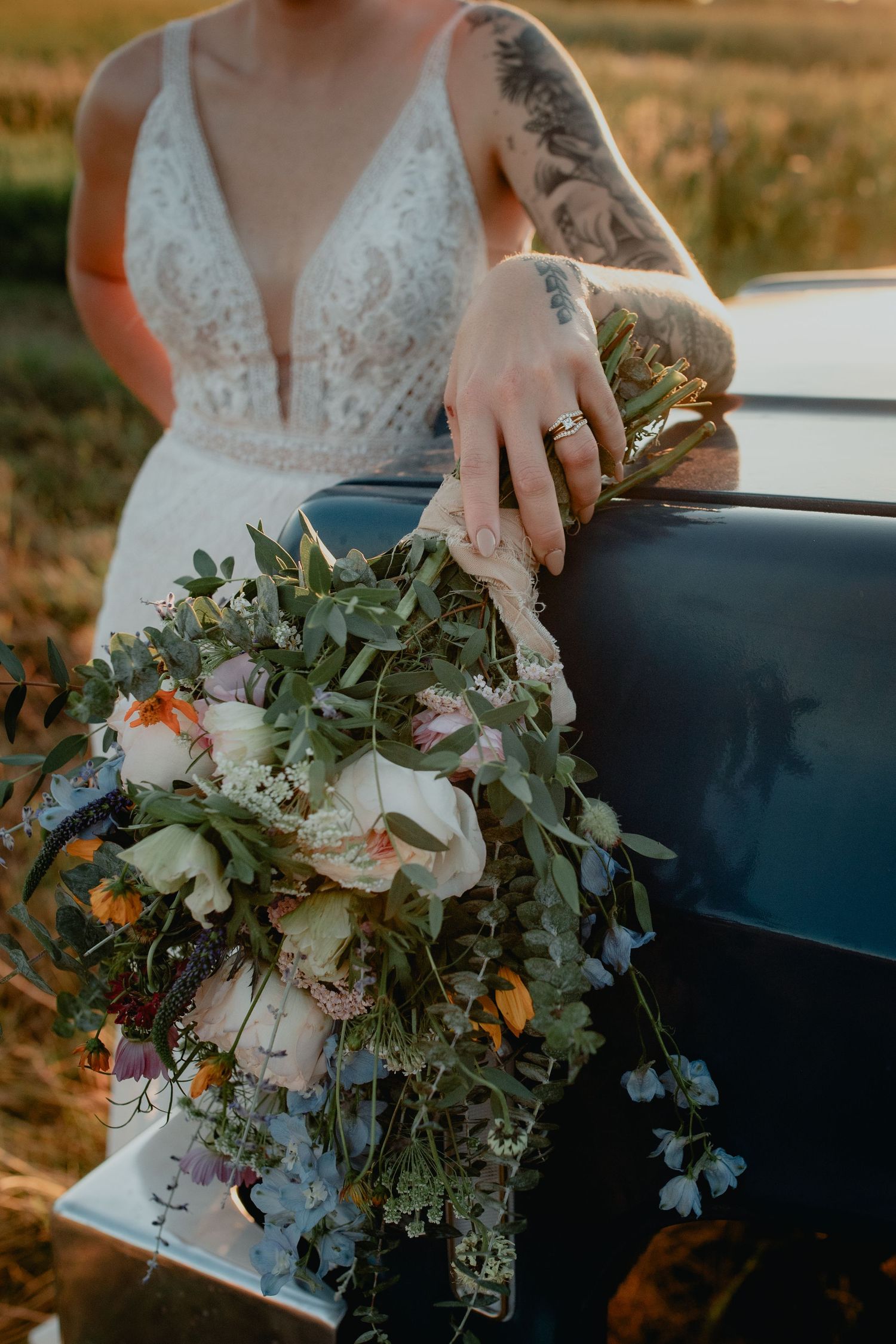 Tattooed bride in lace wedding dress holding cascading wildflower bouquet while leaning on vintage truck at golden hour.