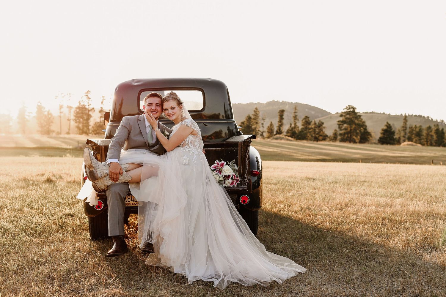 Bride and groom sitting on vintage black truck tailgate in golden field at sunset during outdoor wedding photo session.