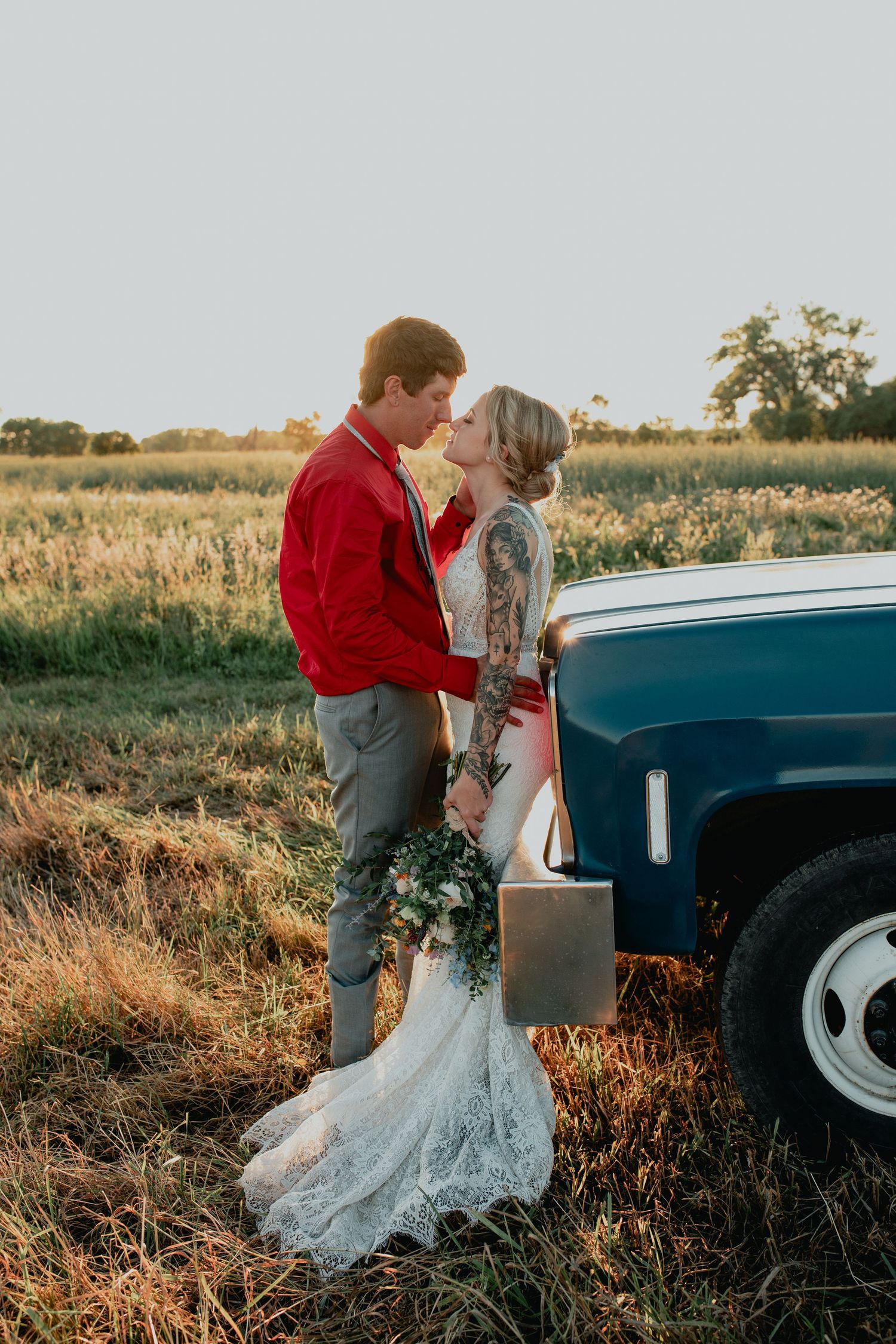 A bride and groom share a romantic moment near a vintage blue truck in a golden field at sunset.