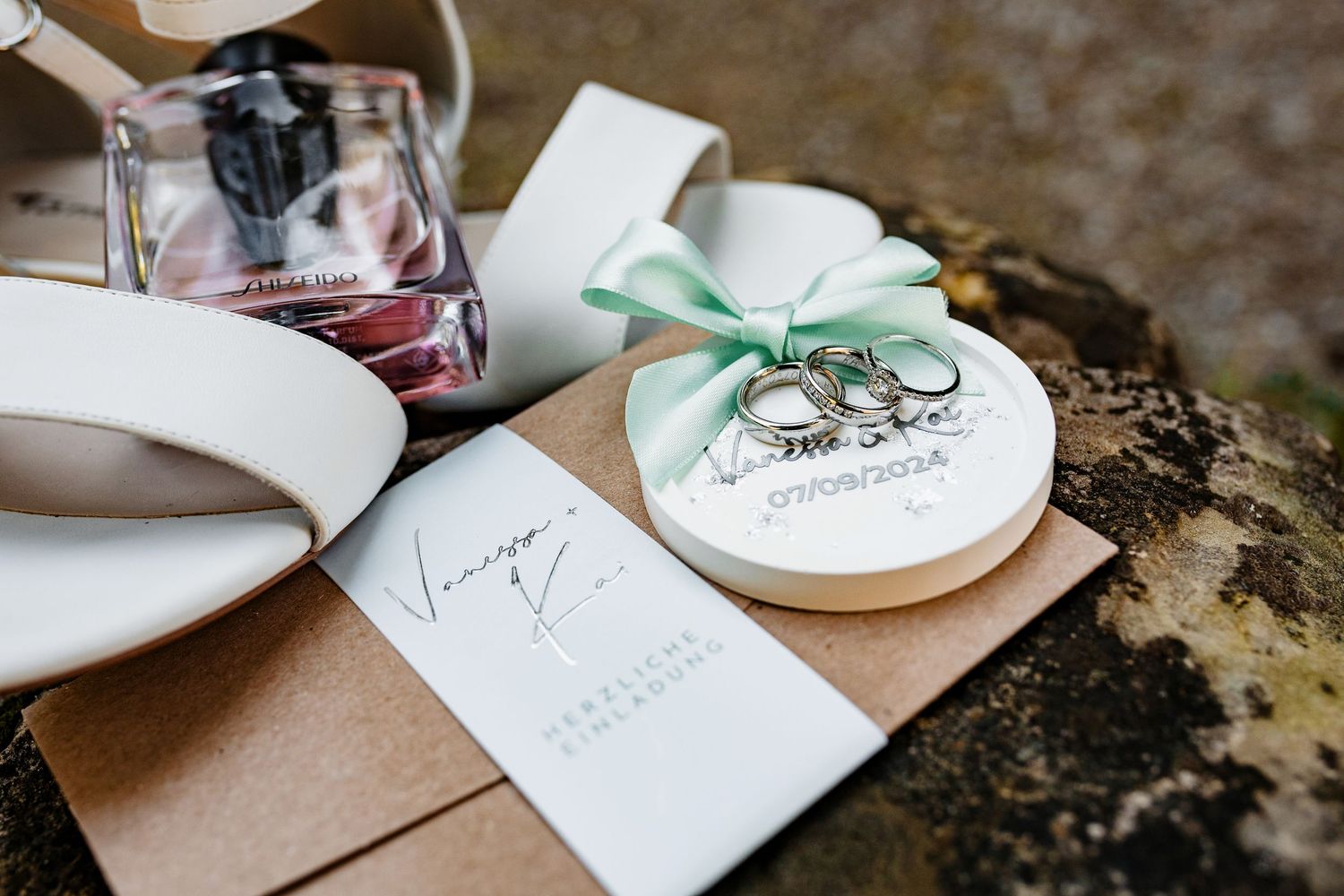 Wedding flat lay with rings on ceramic dish, green ribbon, perfume bottle, and kraft paper invitation on rustic surface.