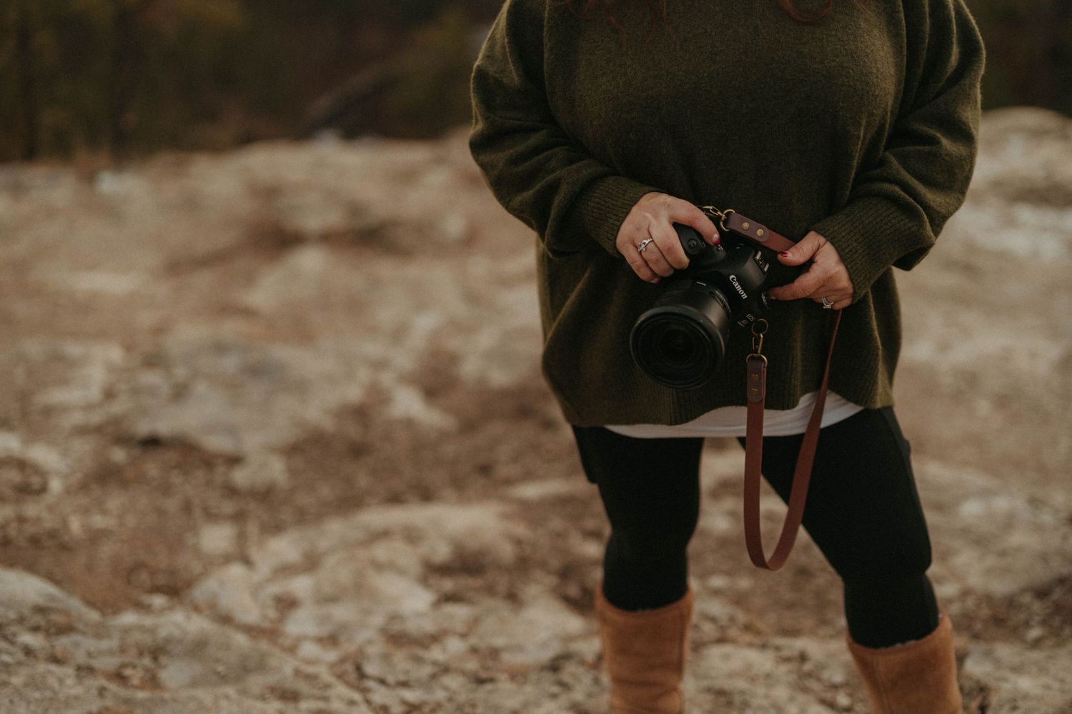 Female photographer in green sweater holding professional DSLR camera with leather strap on rocky outdoor terrain.