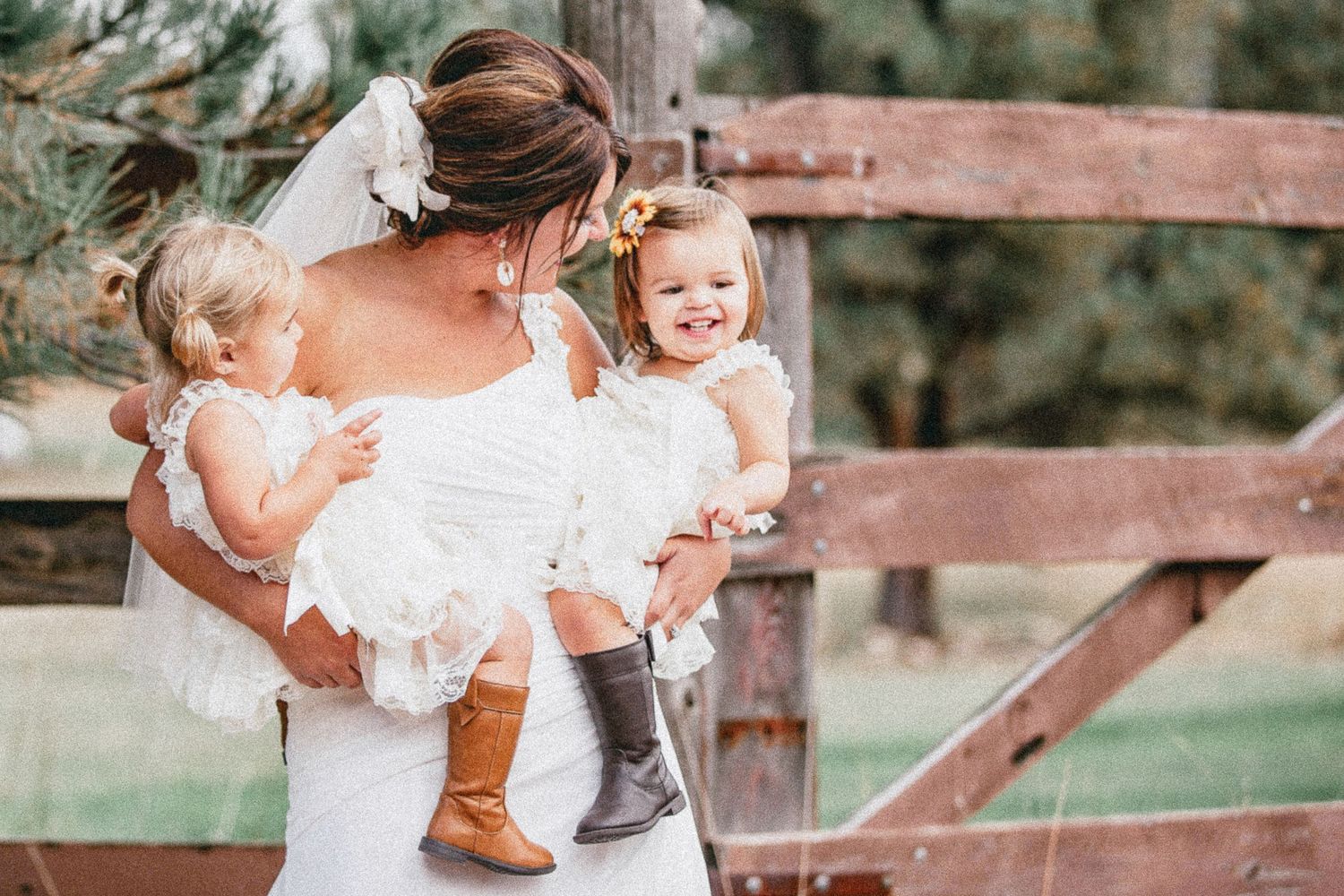 Bride in white dress holding two toddler flower girls in matching white dresses and boots at rustic outdoor wedding.
