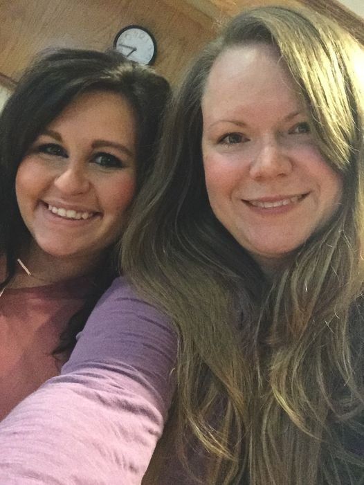 Two smiling women take a selfie together indoors in a warmly lit room.