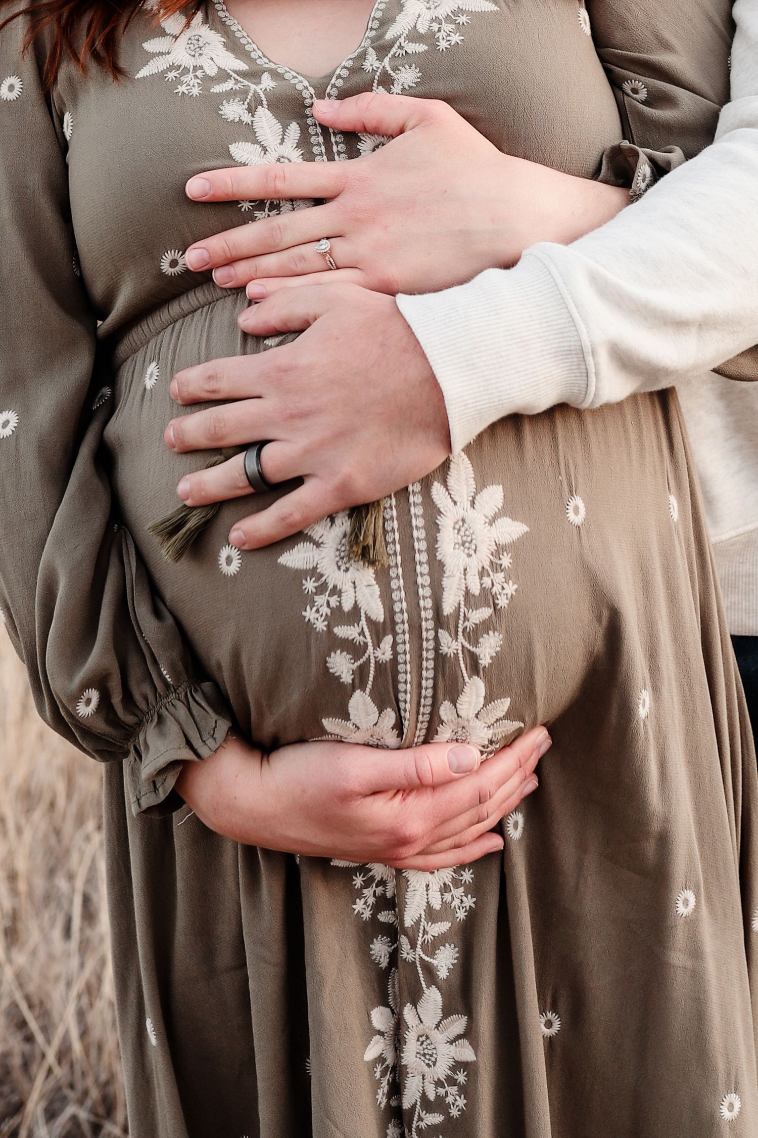 Couple holding pregnant belly together, wearing wedding rings, in floral dress outdoors.