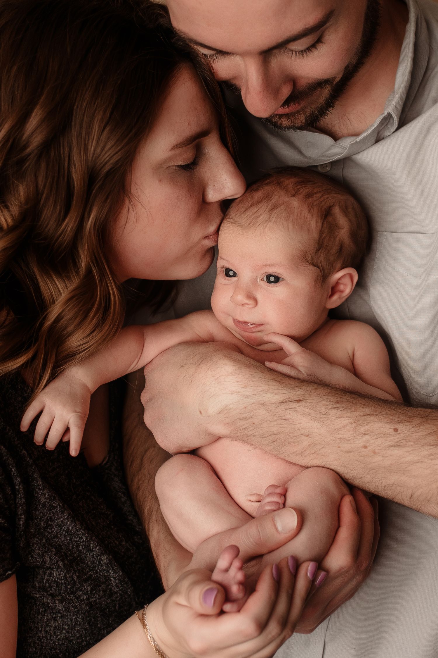 Parents lovingly kissing their alert newborn baby while cradling together in family portrait.