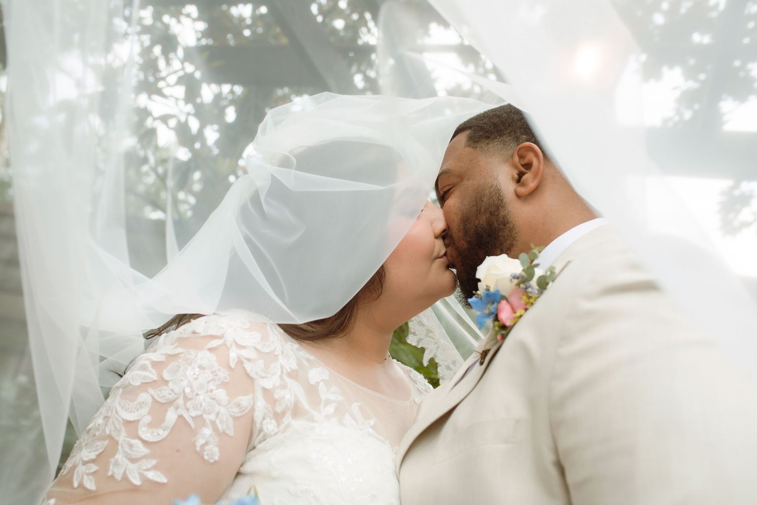 Bride and groom sharing a romantic kiss under a flowing white veil outdoors at their wedding ceremony.