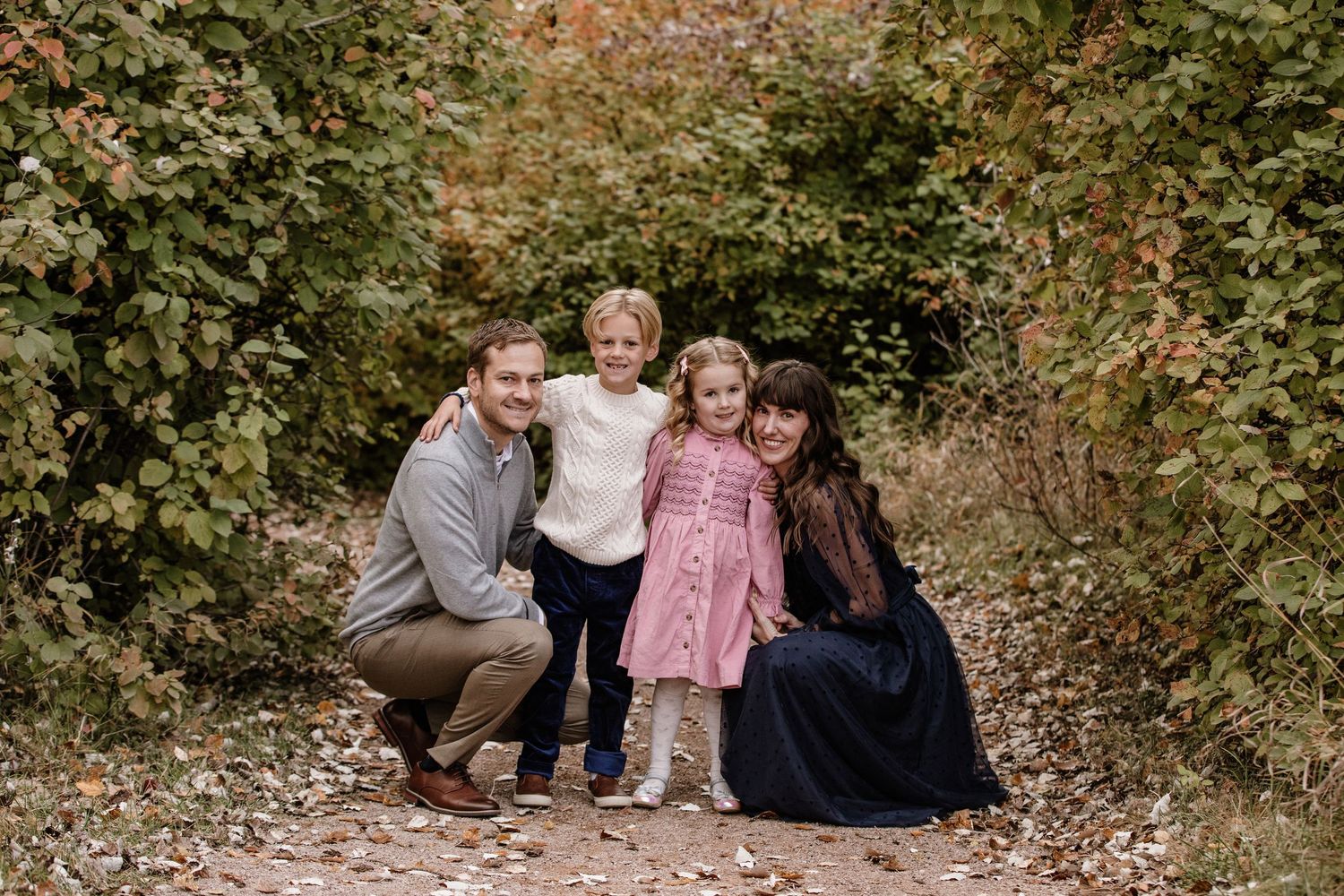 Family of four posing on a forest path surrounded by autumn foliage for a fall outdoor portrait.