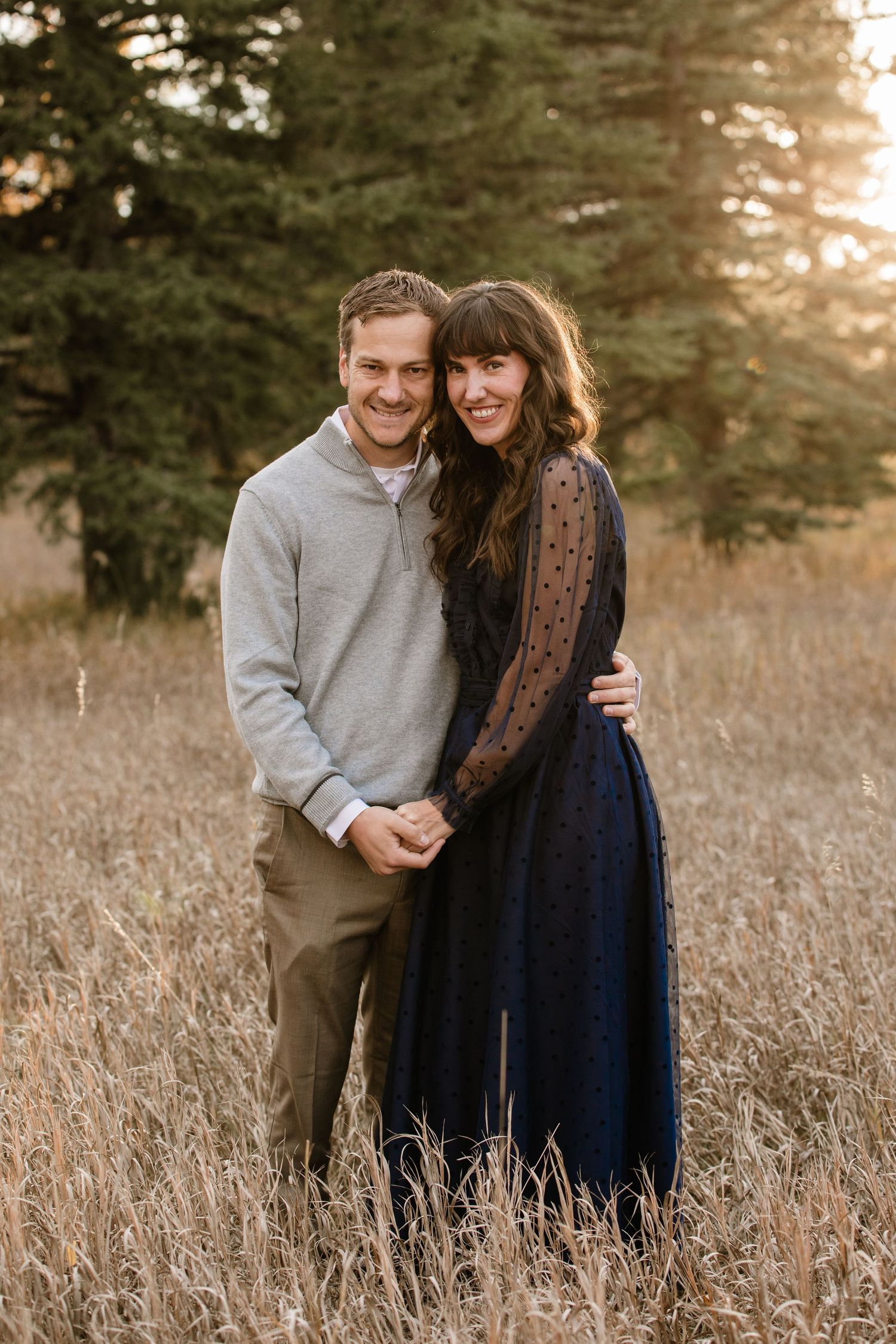 A smiling couple embraces in a golden field, man in gray pullover, woman in navy lace dress, with trees behind.