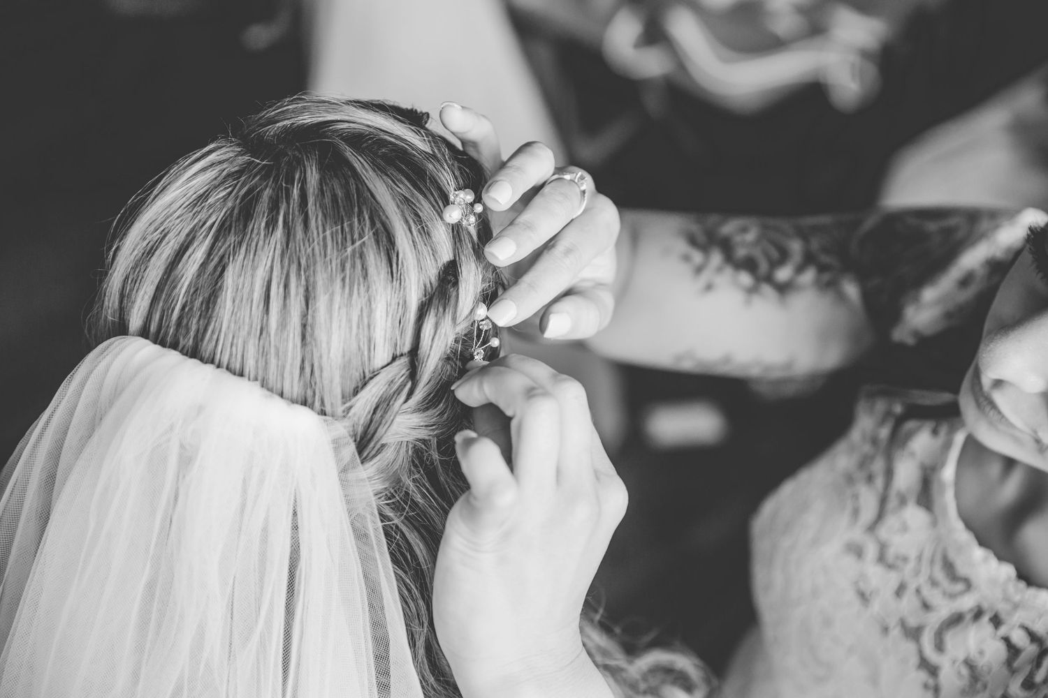 Black and white photo of a bride having her braided hair adorned with flowers by a tattooed bridesmaid.