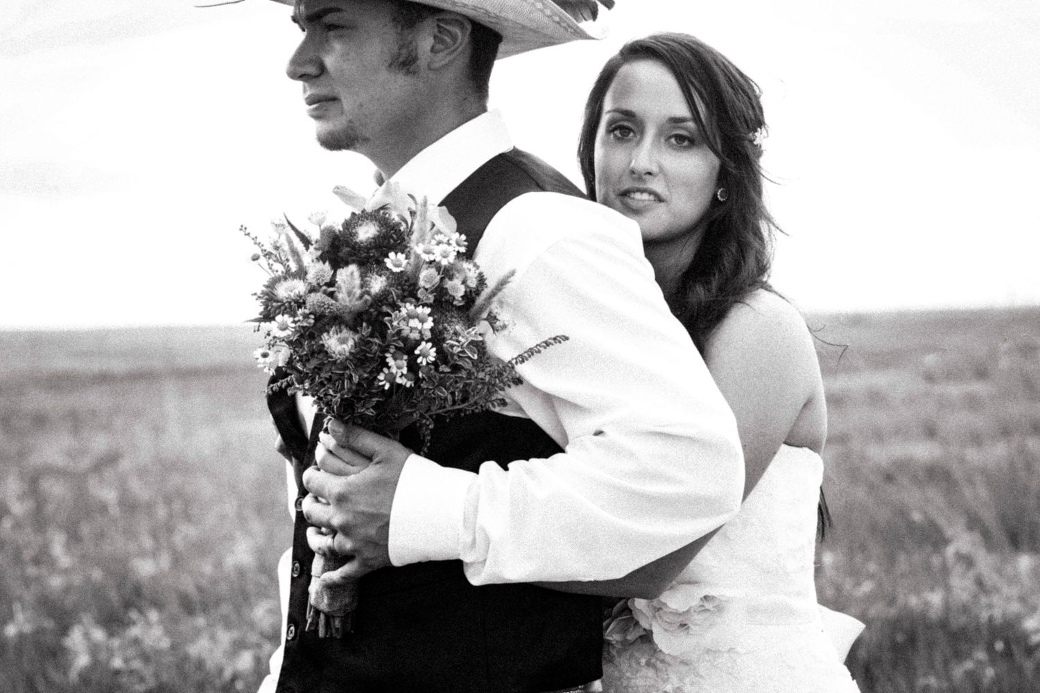 Black and white wedding photo of bride hugging groom in cowboy hat while holding wildflower bouquet in a field.