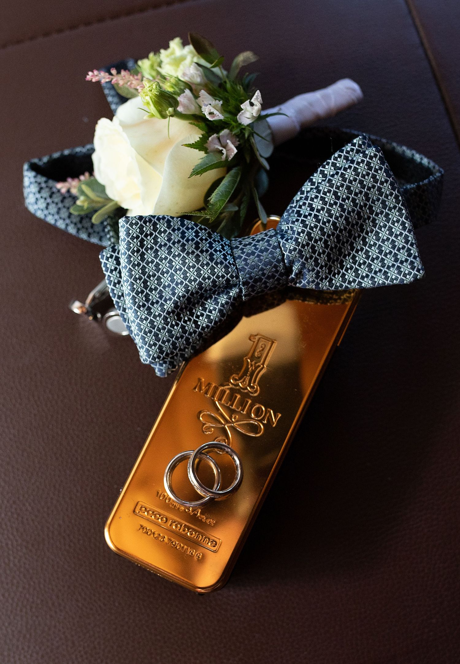 Wedding rings on gold bar beside blue bow tie and white rose boutonniere on brown leather surface.