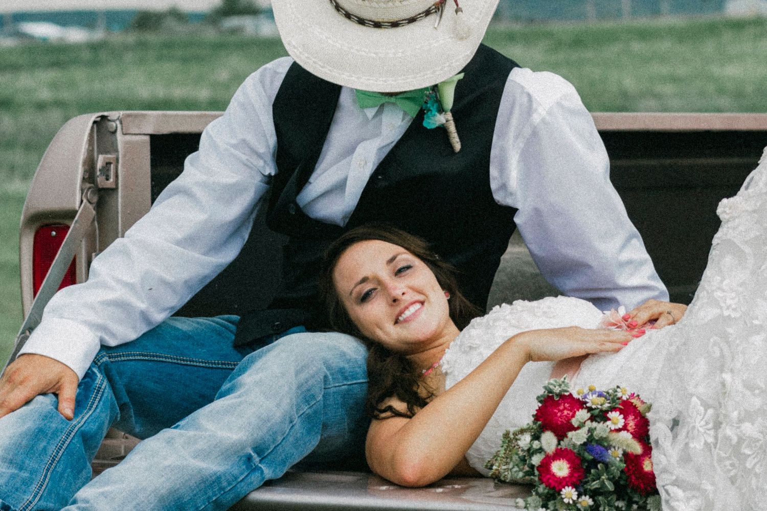 Bride in jeans smiling while lying in truck bed with groom in cowboy hat holding bouquet at rustic wedding.