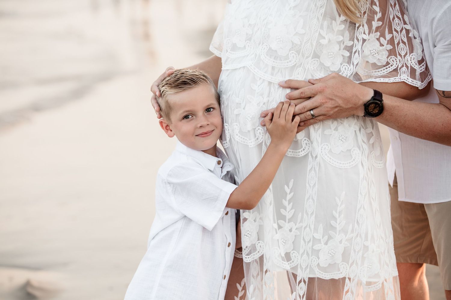 Young boy in white shirt hugging pregnant mothers belly at the beach.