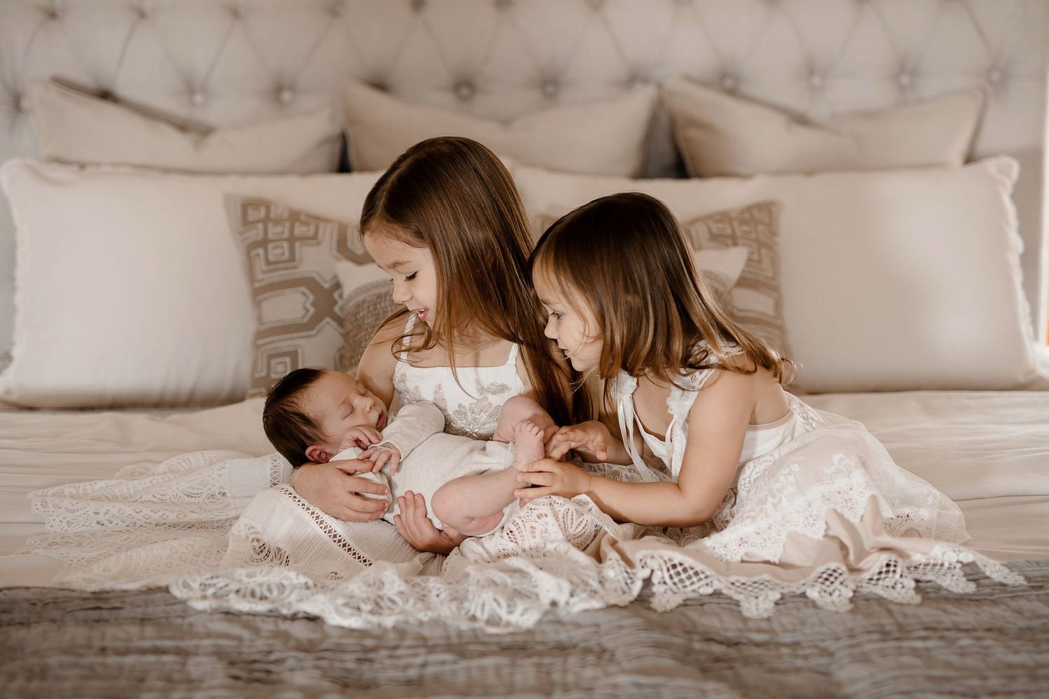 Two older sisters in white dresses sit on a bed admiring their newborn baby sibling.