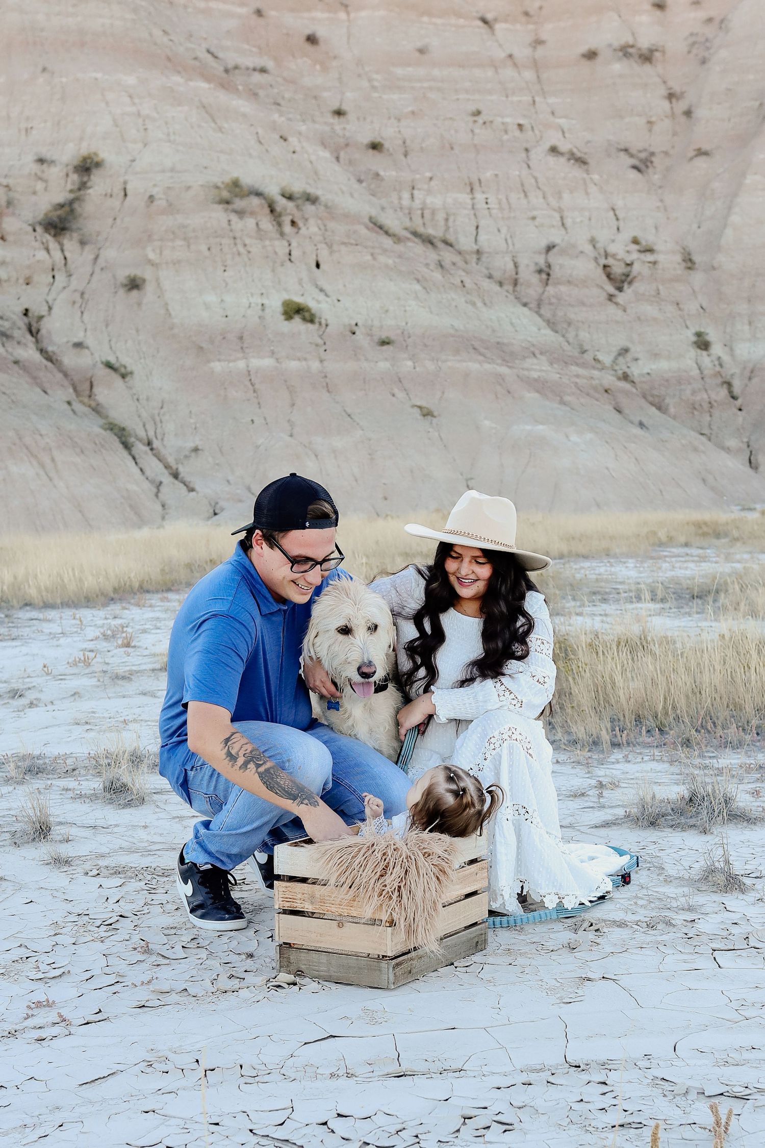Smiling couple with fluffy white dog and baby in wooden crate during desert photo session.