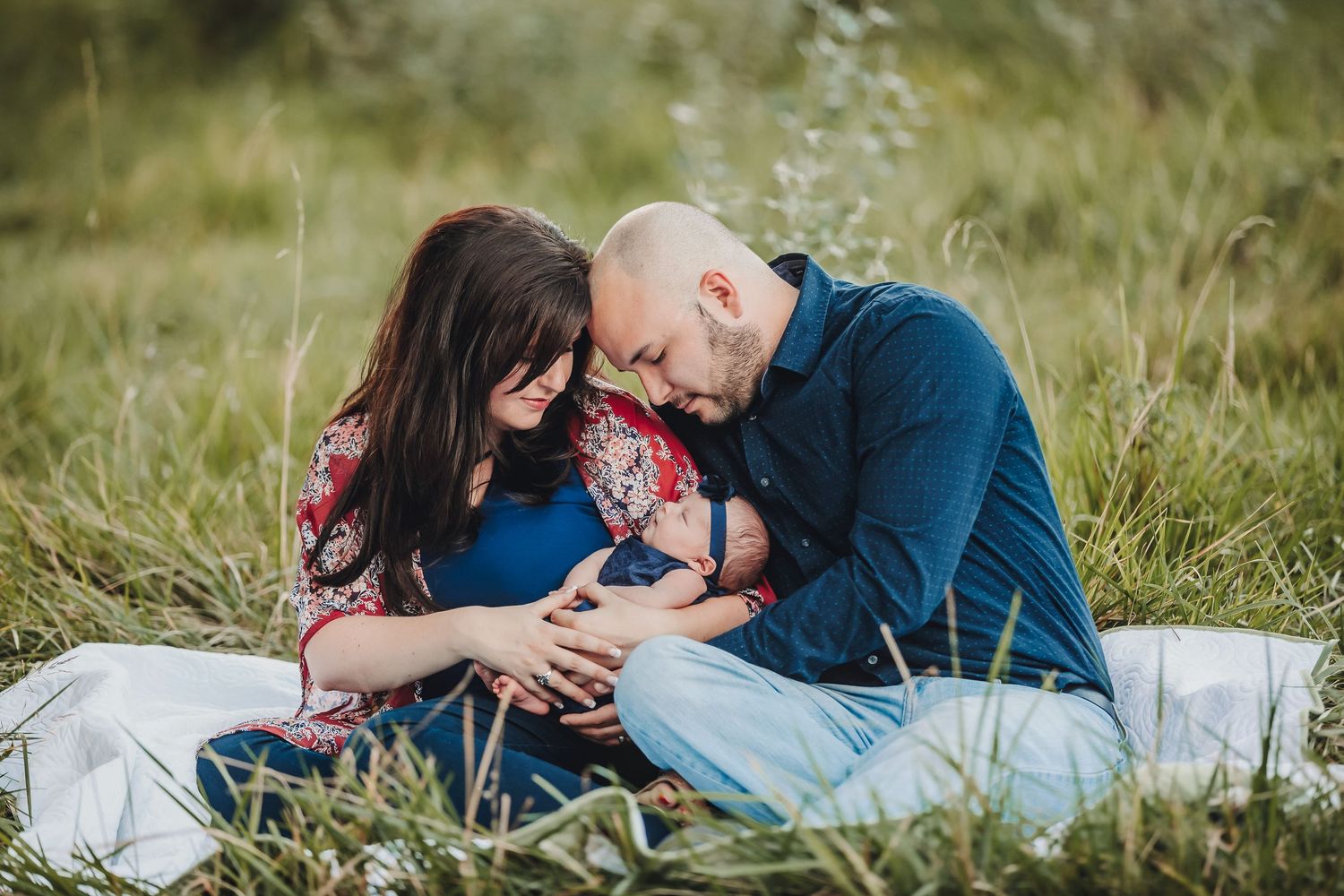 Couple sitting in green grass field, tenderly cradling their newborn baby together outdoors.