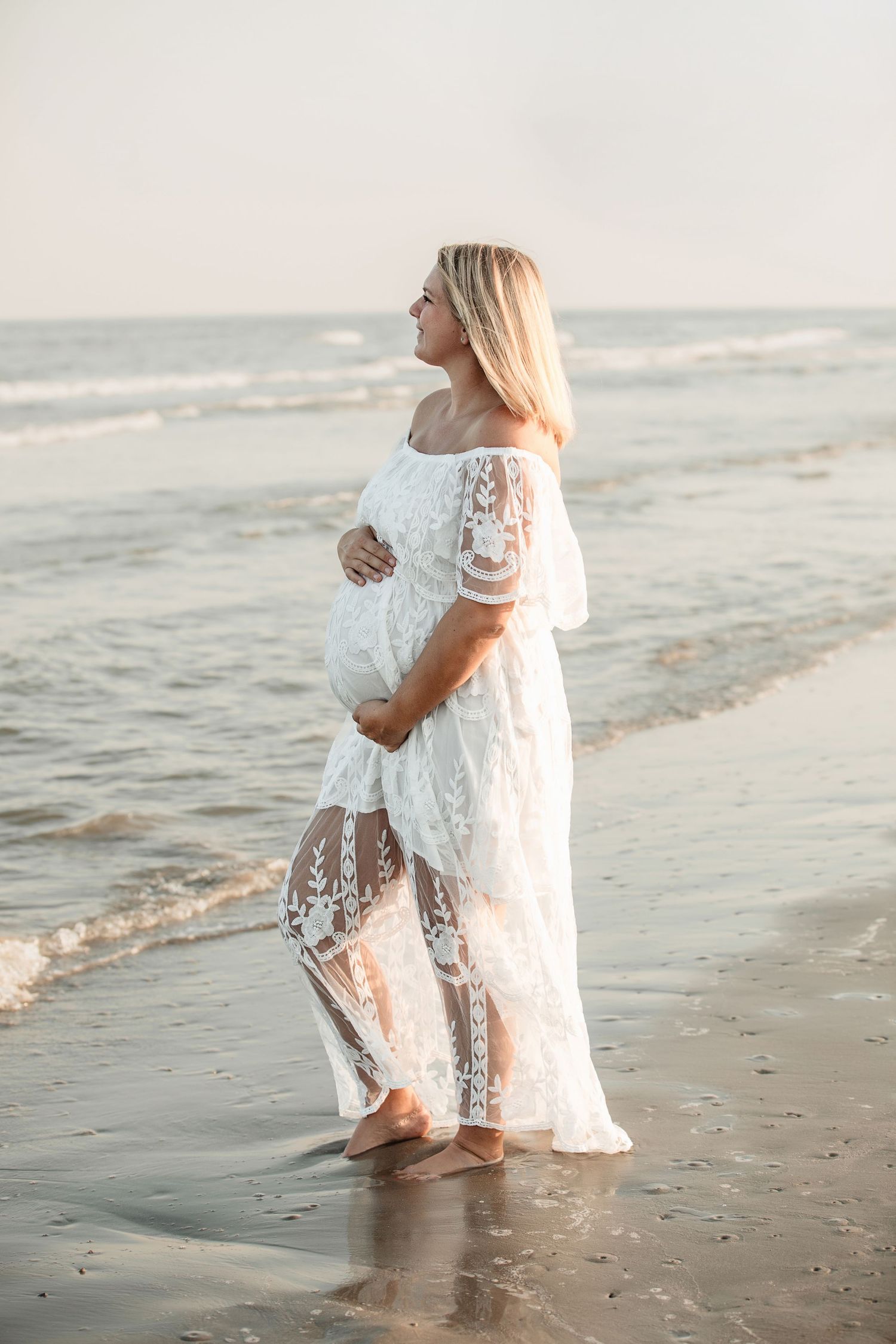 Pregnant woman in white lace off-shoulder gown cradling baby bump while standing in ocean waves at sunset.
