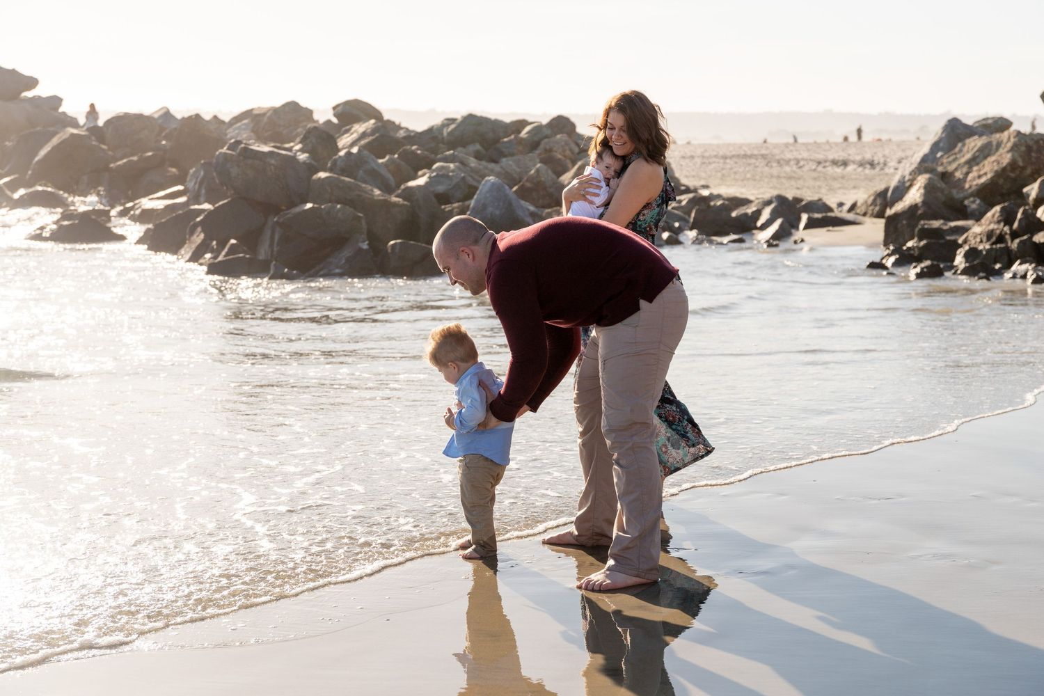 Family with toddler playing at the beach shoreline with rocks in the background on a sunny day.