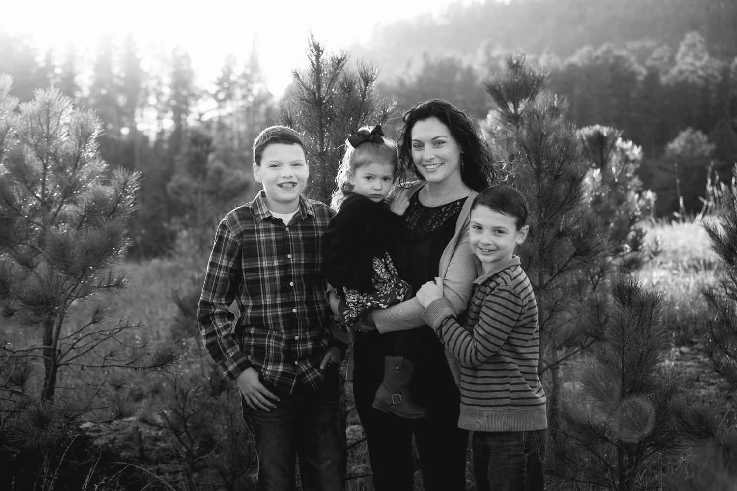 Black and white family portrait of a mother with three children among pine trees.