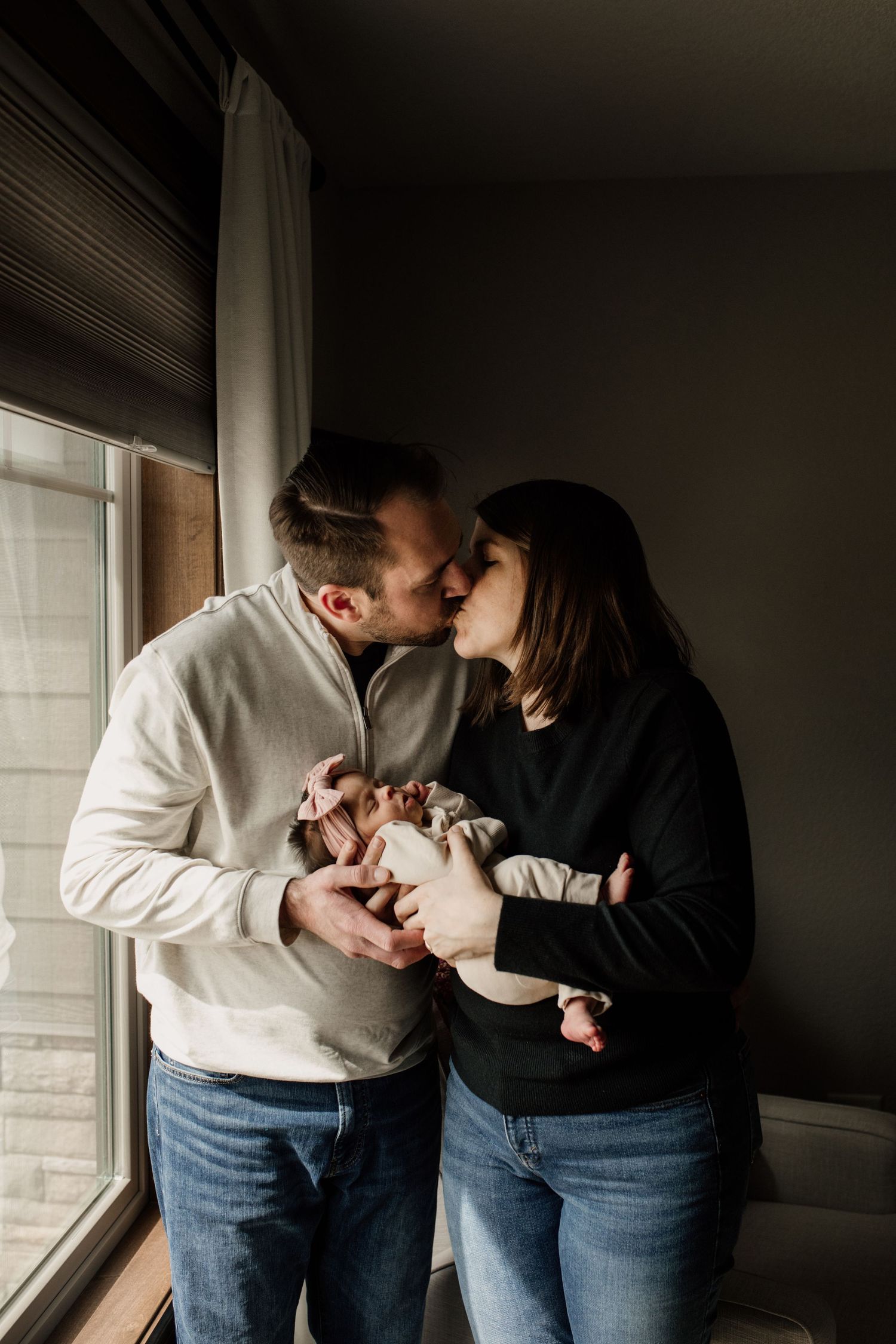 New parents sharing a kiss by a window while gently holding their swaddled newborn baby together.