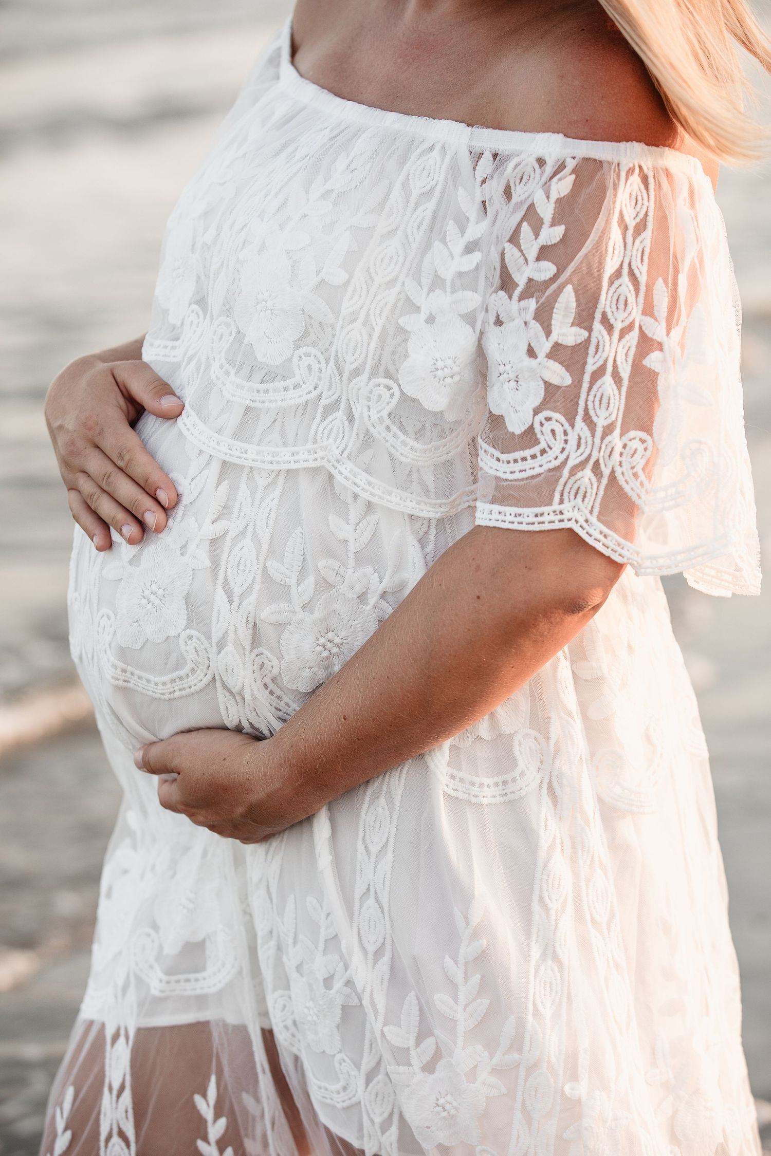 Close-up of pregnant woman in white floral lace dress gently holding baby bump with both hands outdoors.