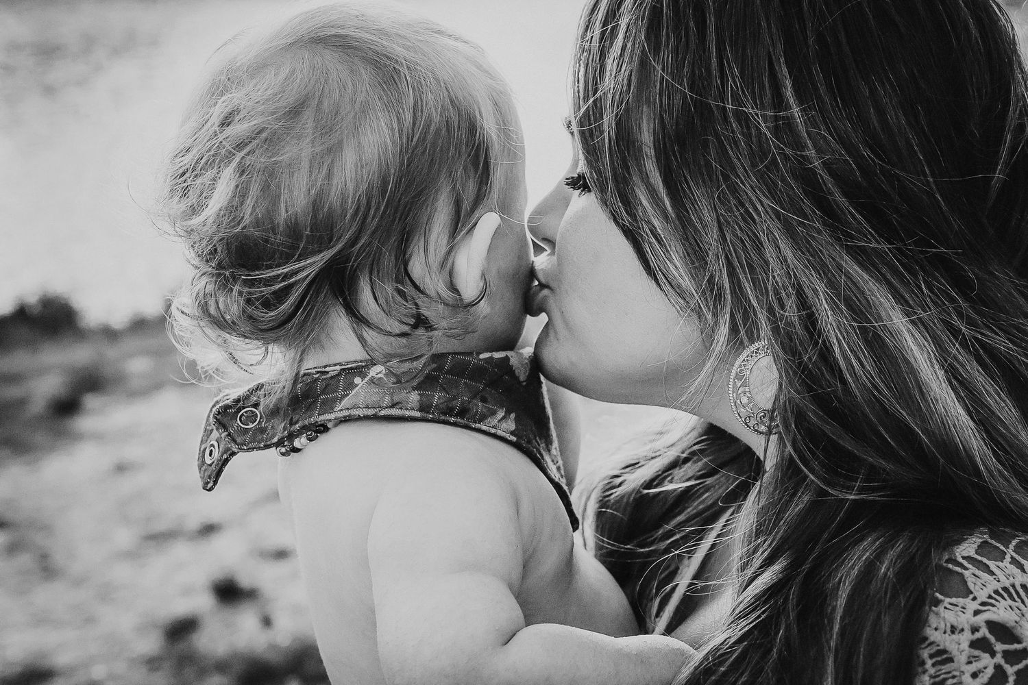 Black and white photo of a mother tenderly kissing her baby outdoors.