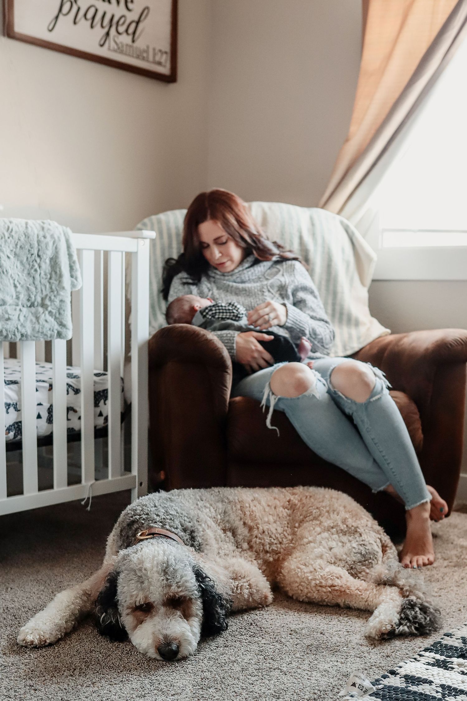 Mother in gray sweater holds newborn in nursery chair while large fluffy dog rests on carpet beside white crib.