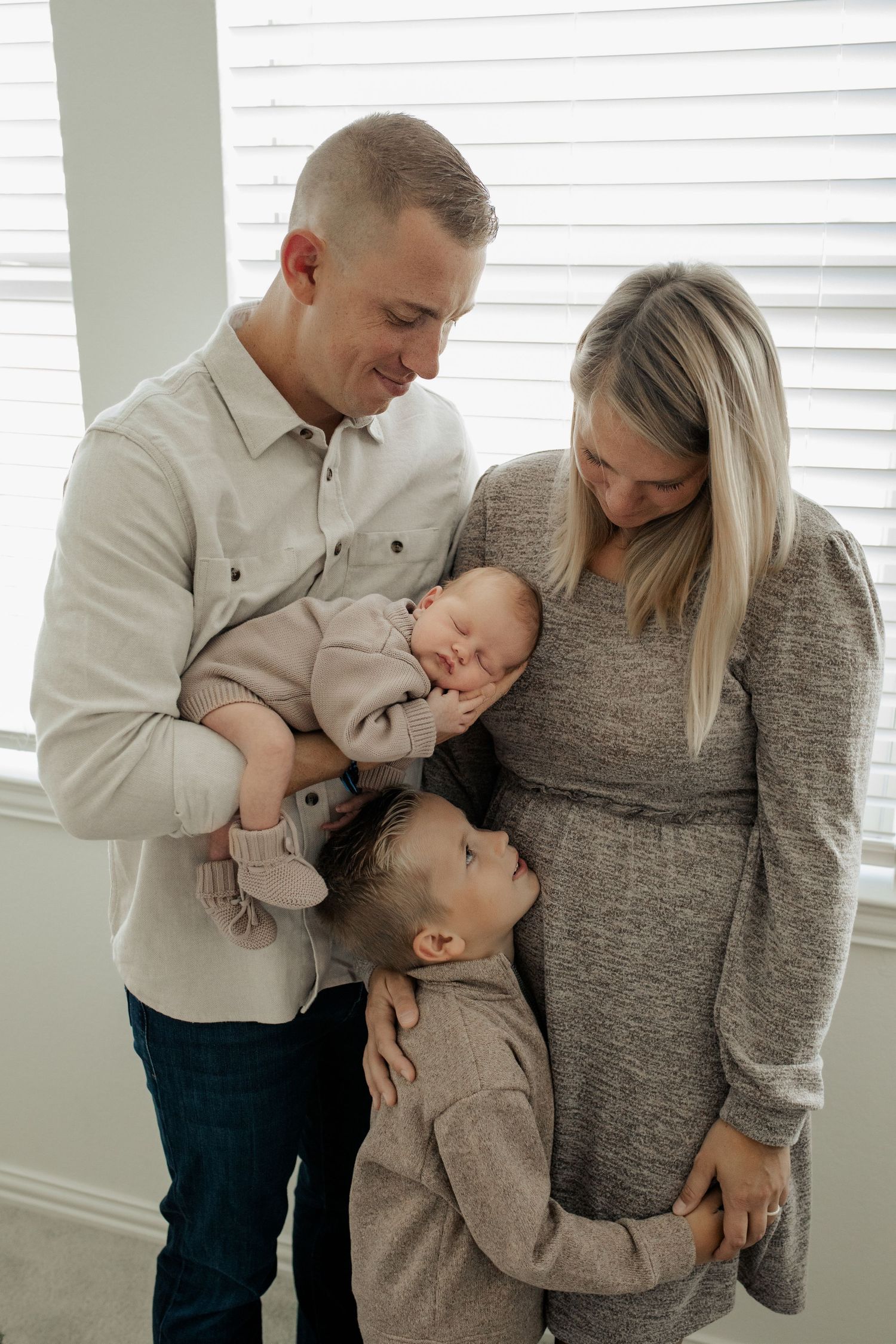 Family of four with father holding newborn baby while young son hugs mother in bright white room.