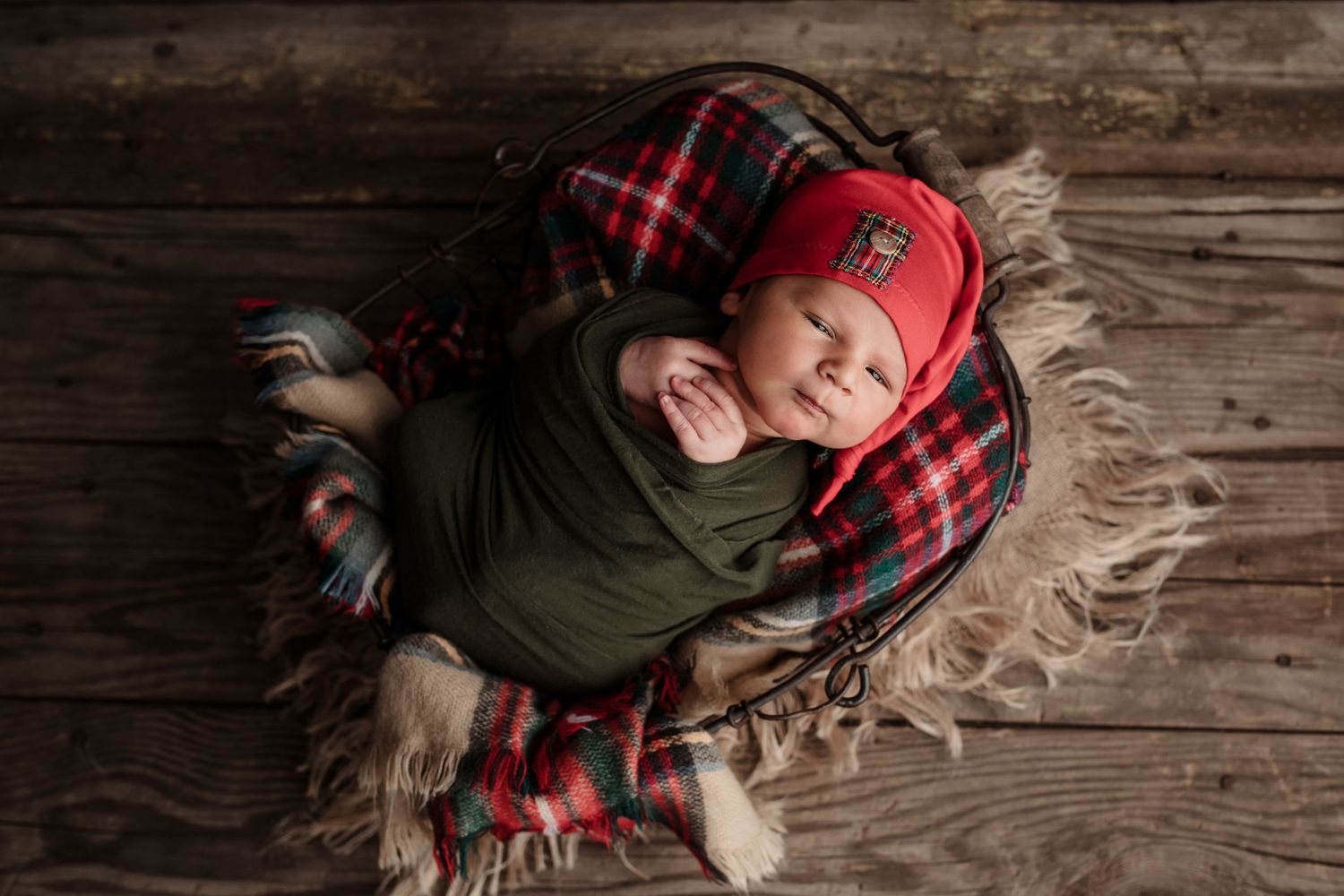 Sleeping newborn baby swaddled in green wrap wearing red plaid cap in rustic basket.
