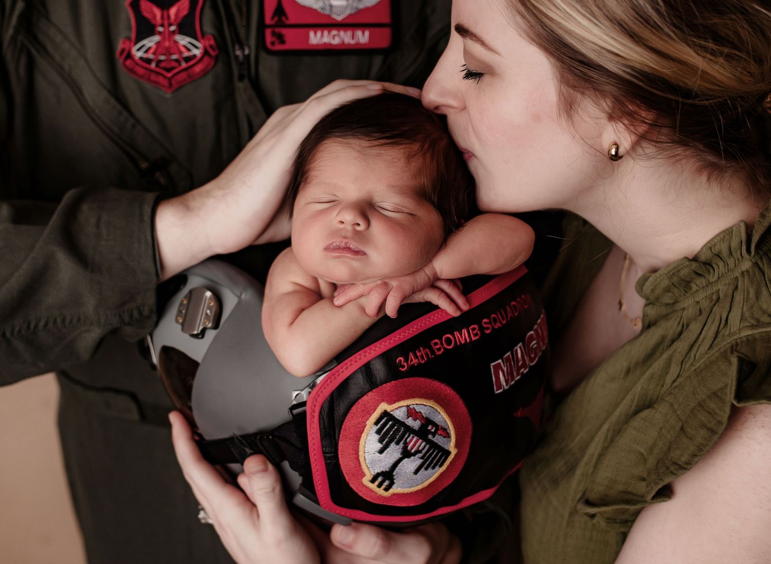 Parents in military flight suits lovingly kissing newborn baby resting on helmet.