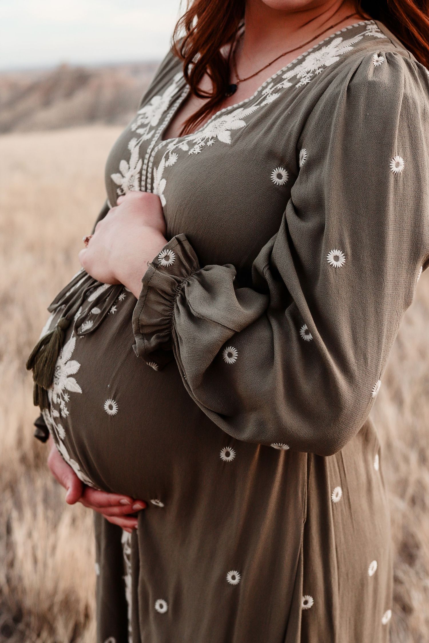 Pregnant woman in olive floral embroidered dress cradling baby bump outdoors.