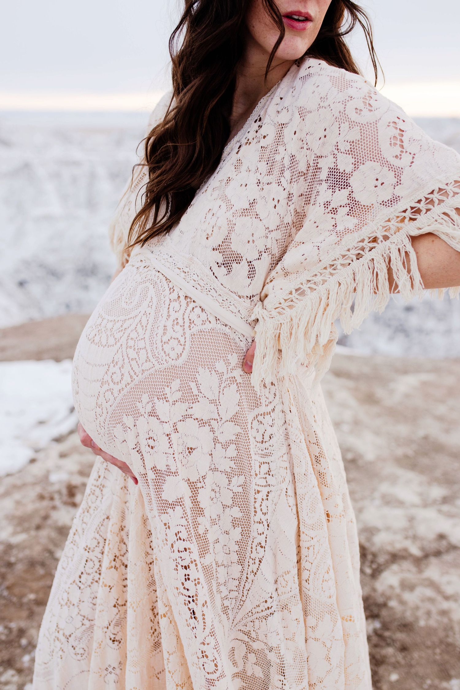Close-up of pregnant woman in cream boho lace fringe dress cradling baby bump outdoors.