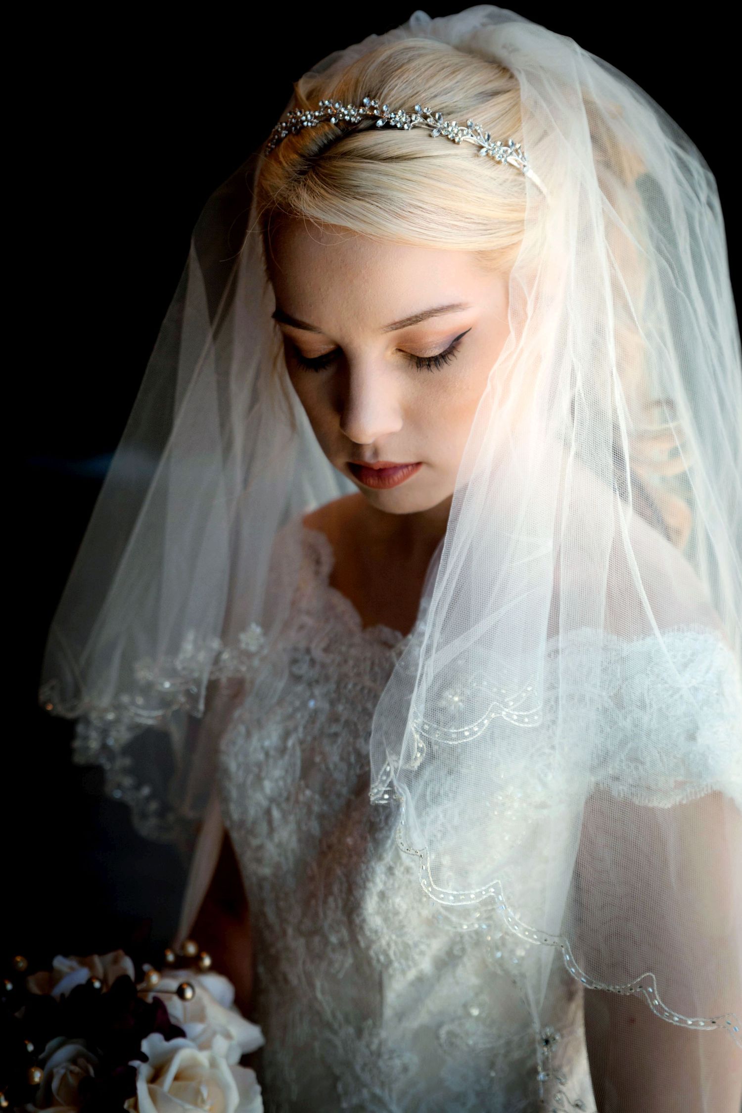 Elegant bride in lace wedding dress and veil with crystal tiara, looking down holding a bouquet.