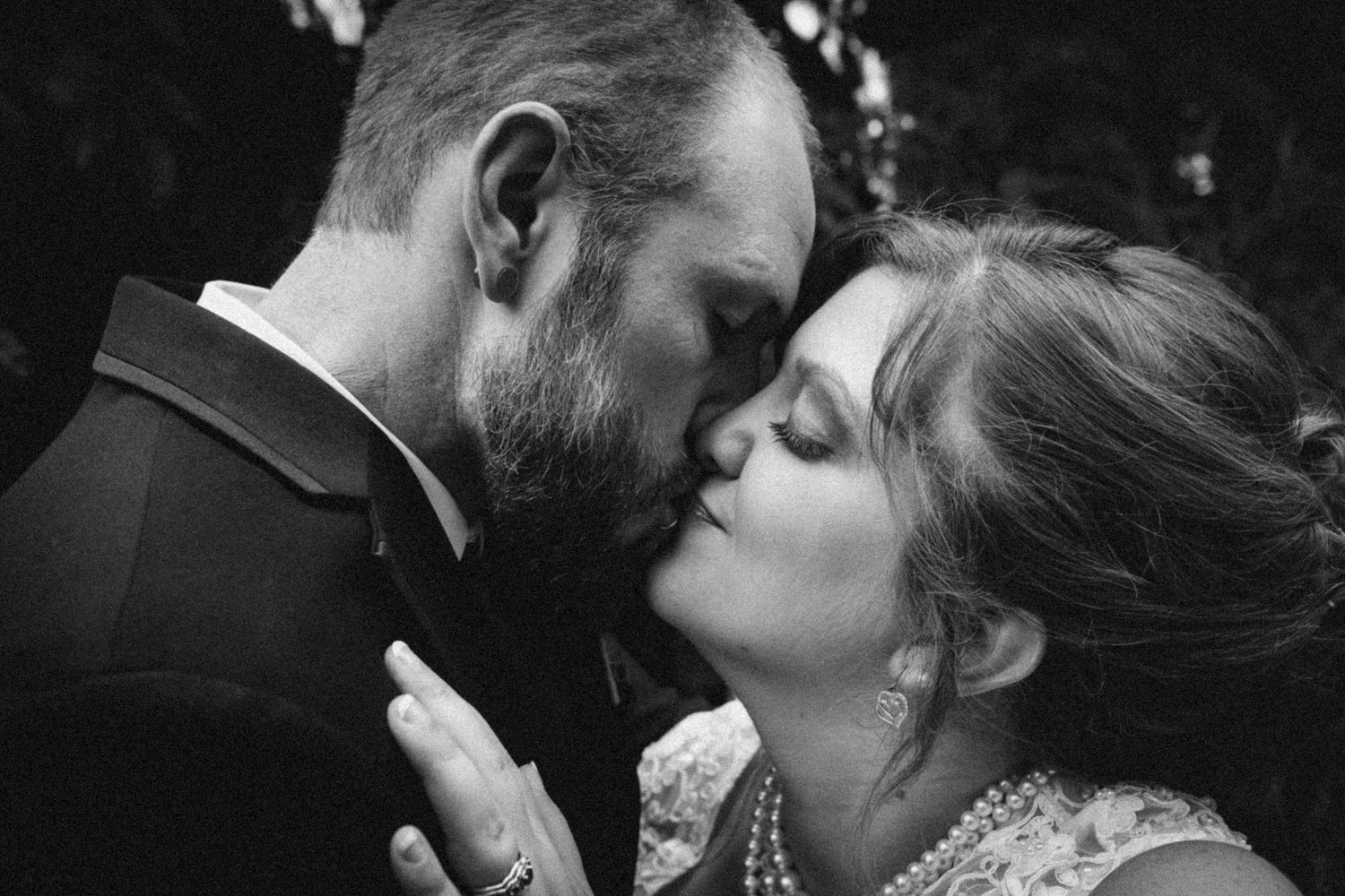 Black and white photo of a couple sharing a romantic kiss, bride wearing pearl necklace and lace dress on wedding day.