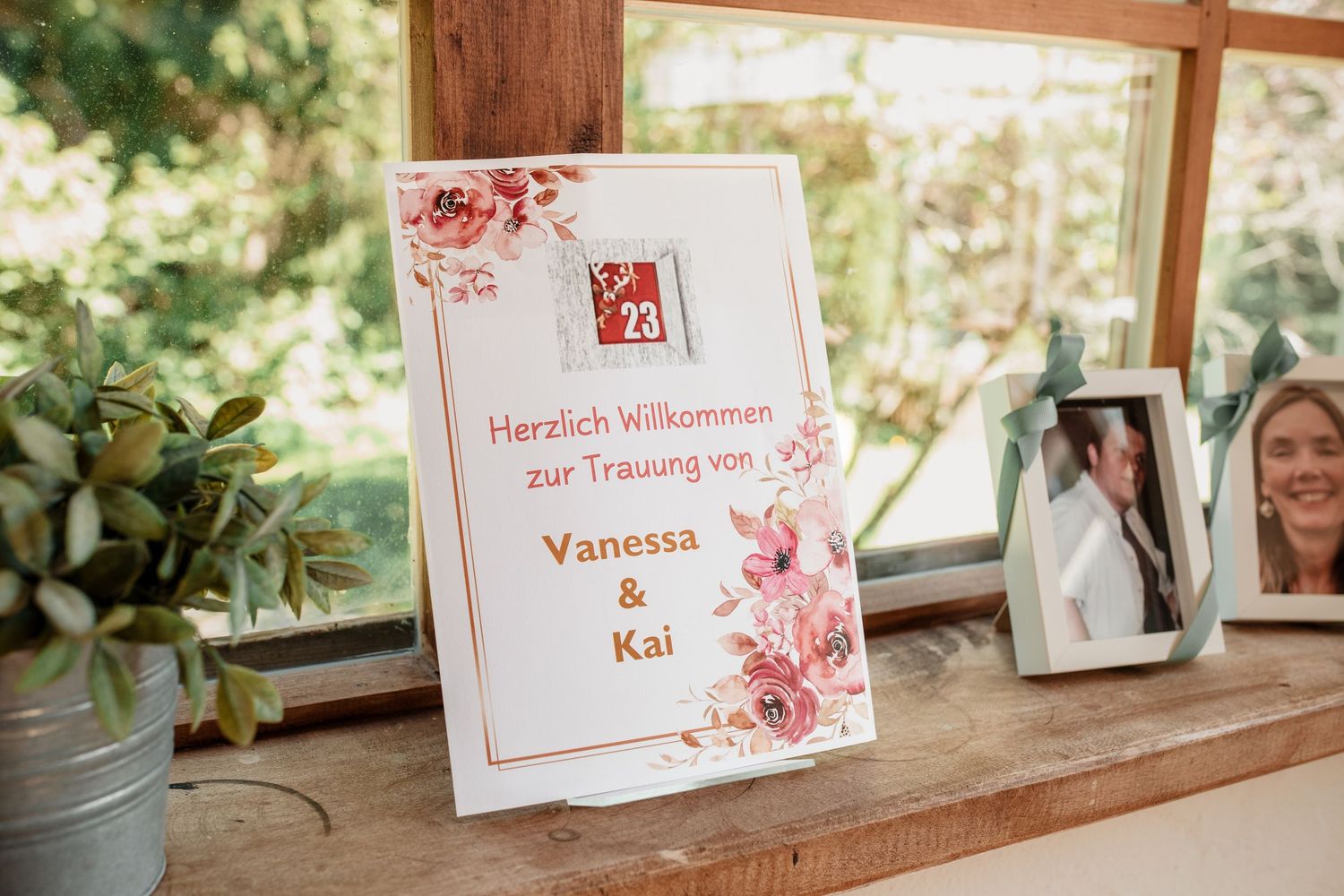 Wedding welcome sign in German for Vanessa and Kai's ceremony, decorated with pink roses on a rustic wooden shelf.