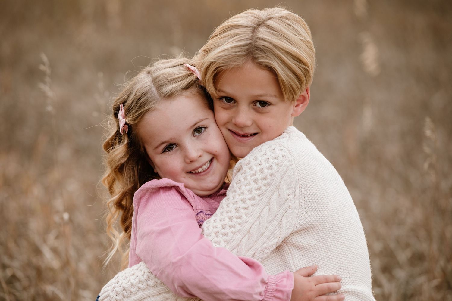 Two blonde children hugging in a golden field, smiling warmly in a sweet outdoor portrait.