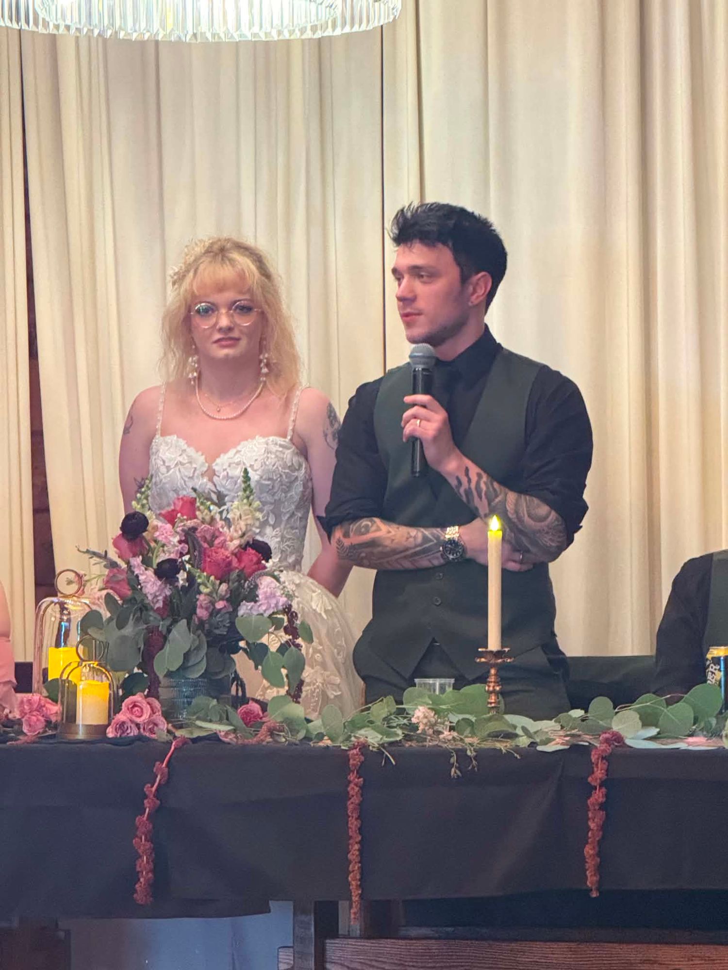 Bride and groom stand at wedding reception head table with floral arrangements, groom speaks into microphone wearing tattooed arms.