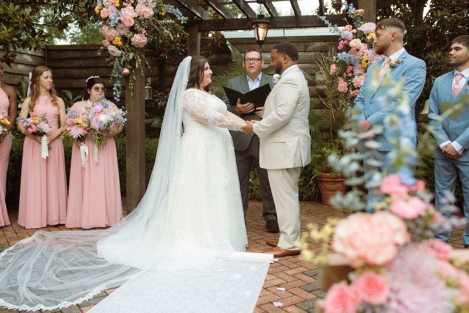 Outdoor wedding ceremony with bride in white gown and veil, groom in tan suit, bridesmaids in pink dresses, floral arch backdrop.