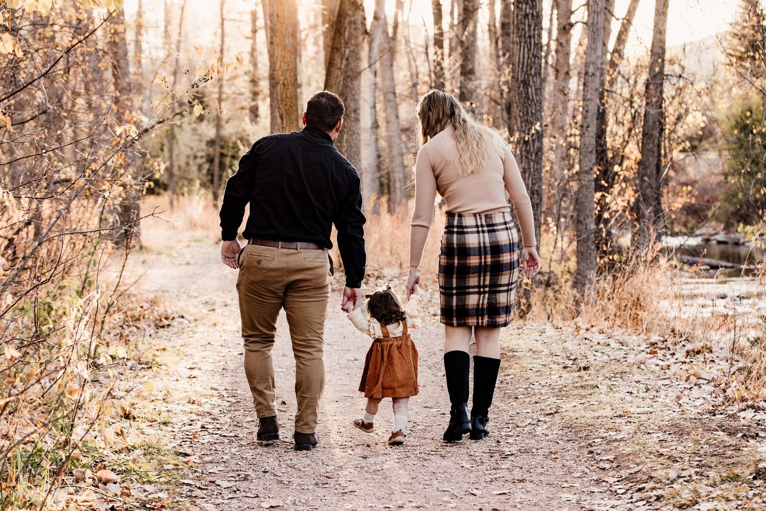 Family of three walking hand-in-hand down a wooded autumn trail, parents holding toddler in fall outfits.