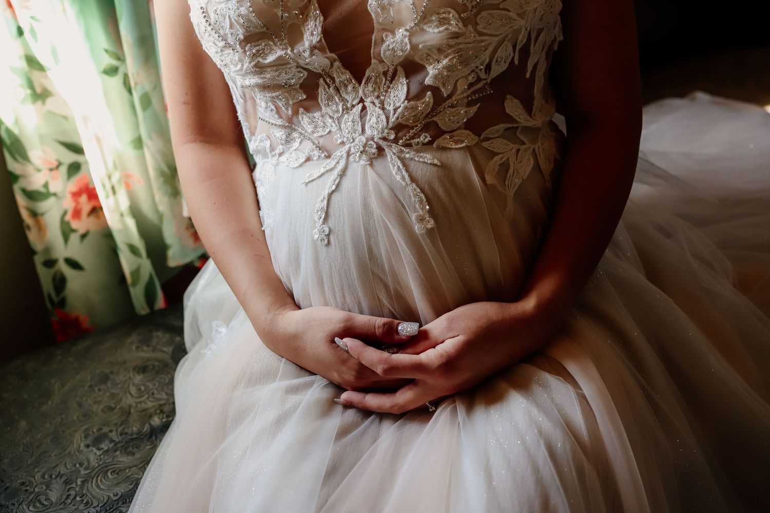 Bride in lace wedding gown with tulle skirt clasping hands, showing intricate floral bodice details.