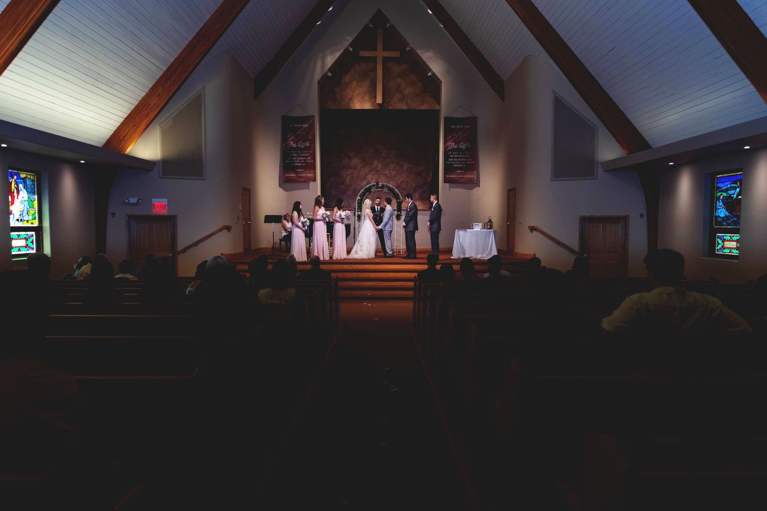 Wedding ceremony in a dimly lit church with bridal party at the altar beneath a cross and stained glass windows.